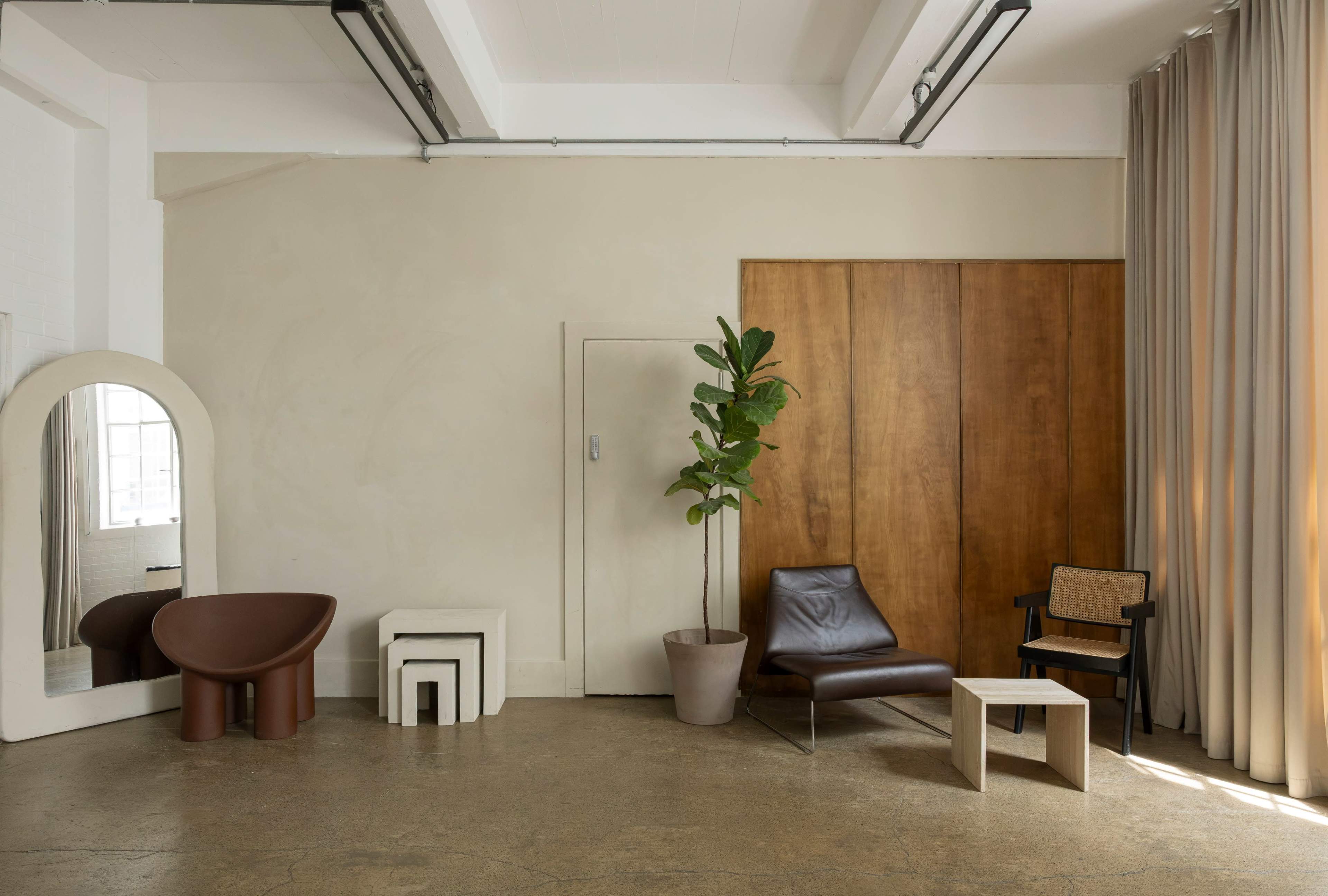 The image shows a minimalist interior space featuring a brown leather chair, a unique brown sculptural table, a small beige table, and a tall plant next to a wall with wooden panels and curtains.