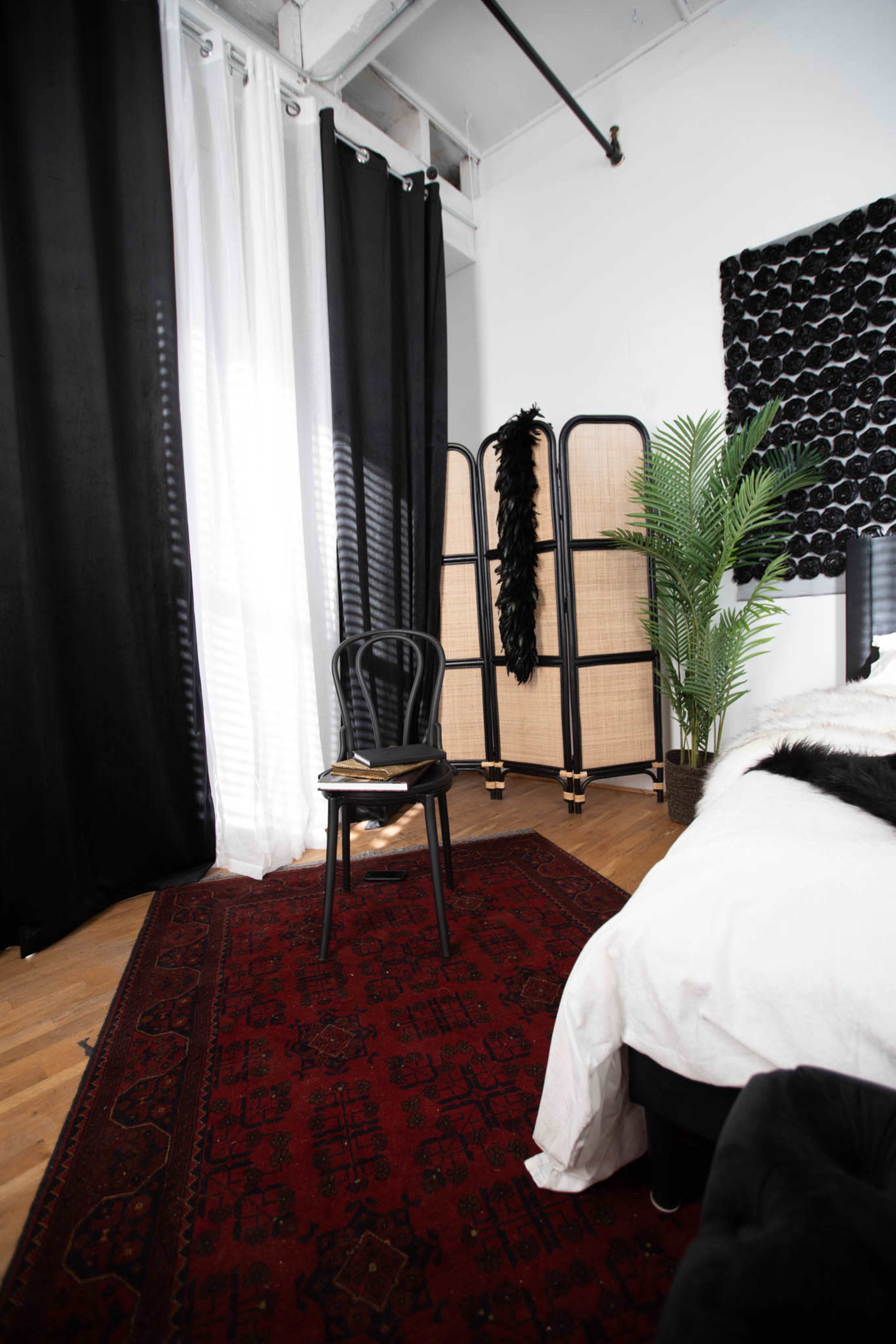 A chair sits on a patterned rug in a room with black and white curtains, a folding screen, and a potted plant.