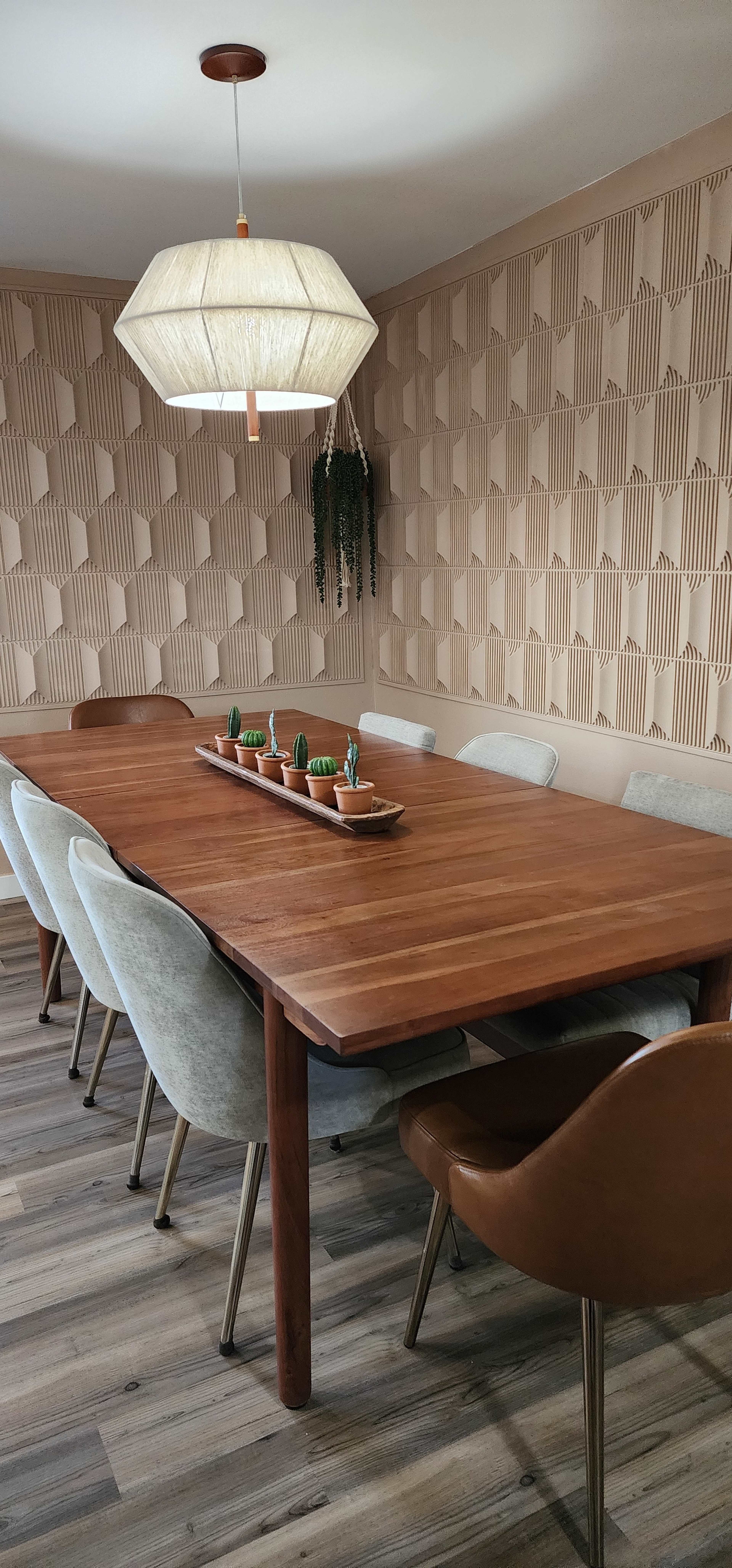 A wooden dining table with six upholstered chairs is set against a textured, patterned wall and features a pendant light above, along with small potted plants in the center.