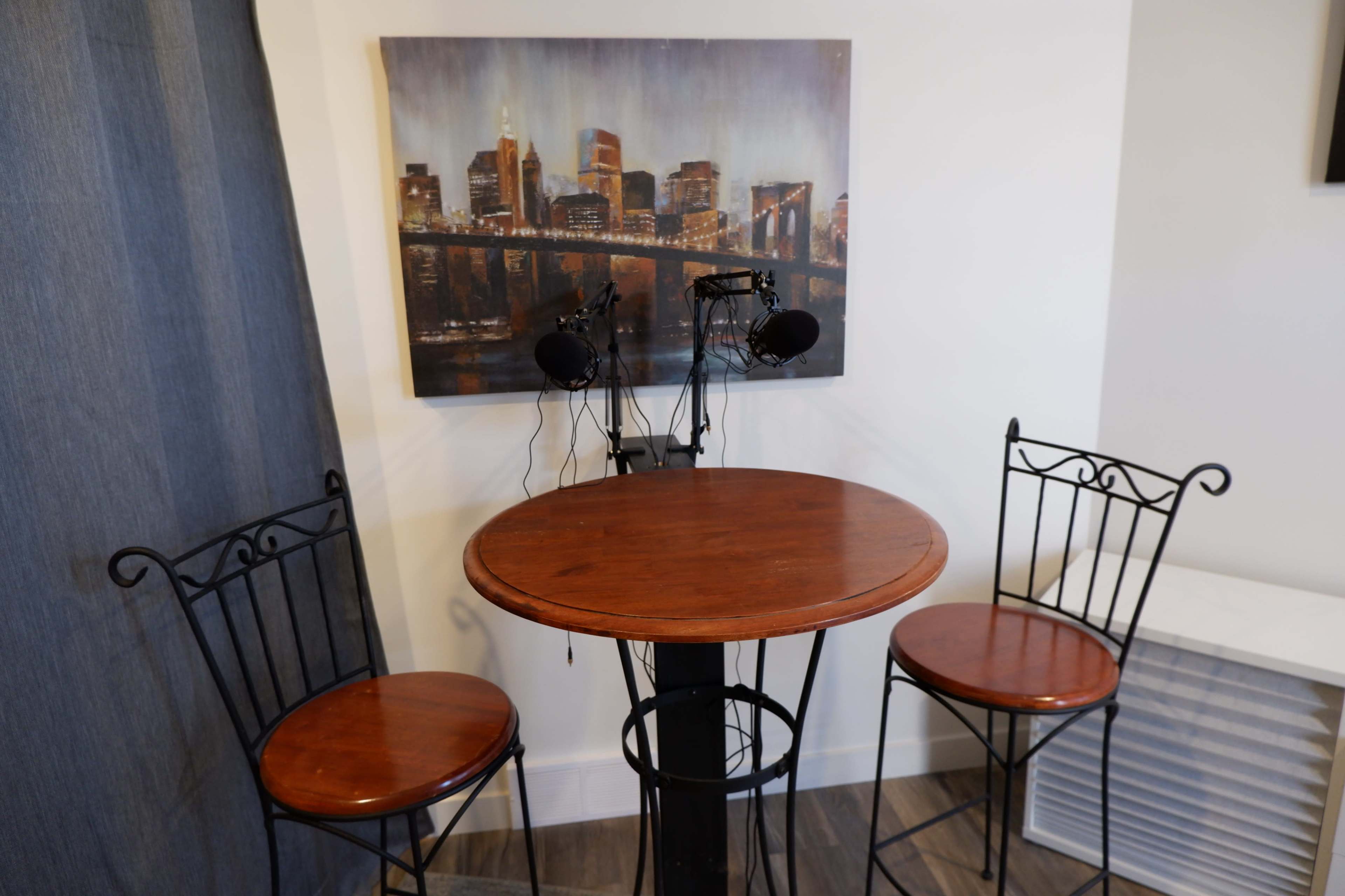 A small round wooden table is set between two metal chairs, with a cityscape painting hanging on the wall above and microphones positioned on the table.