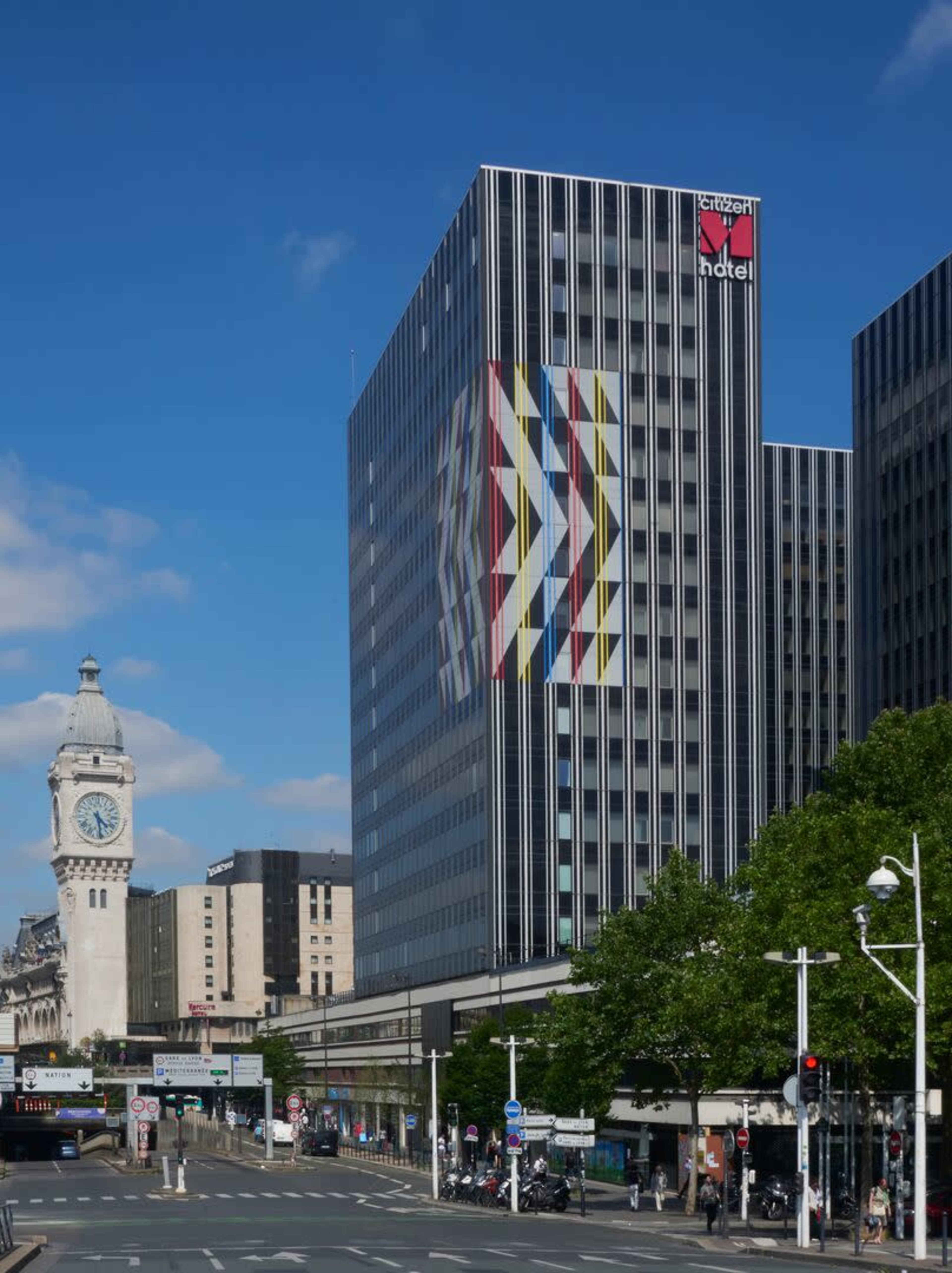 The image shows a large modern building with colorful geometric patterns on its facade next to an older clock tower under a blue sky.