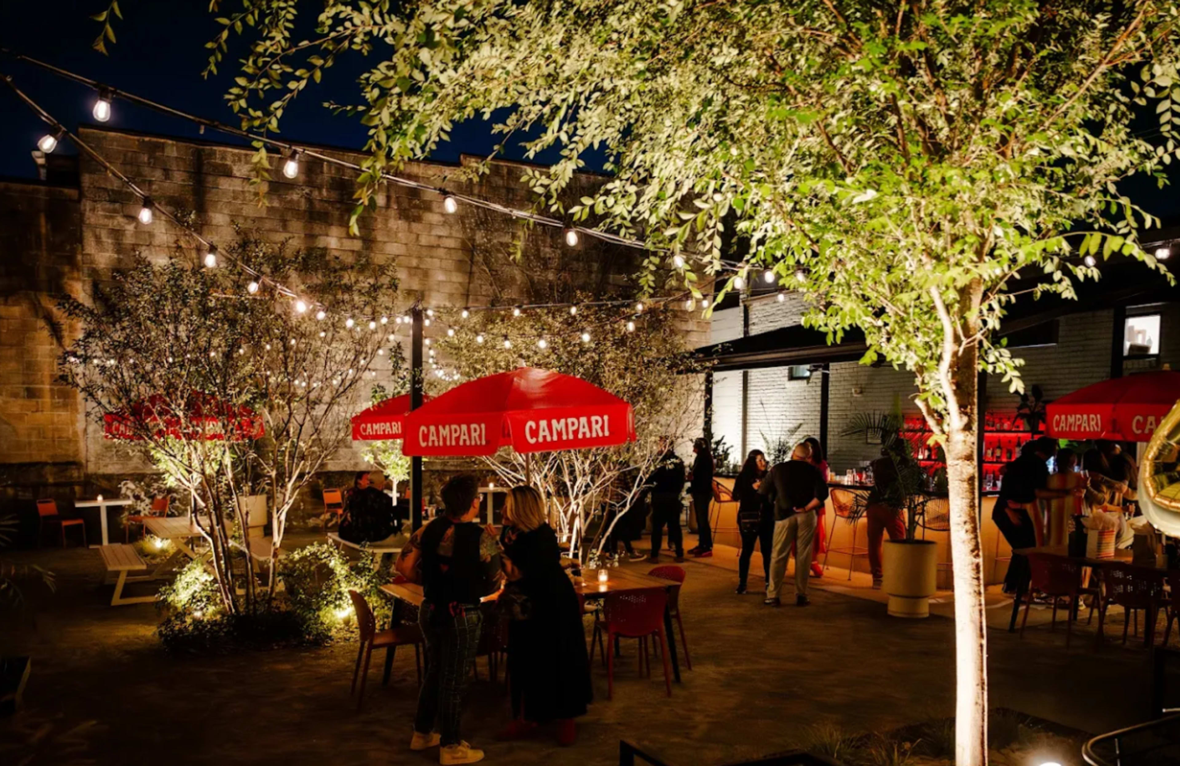 The scene shows a lively outdoor bar area illuminated by string lights, featuring red umbrellas with "Campari" branding and patrons socializing among tables and greenery.