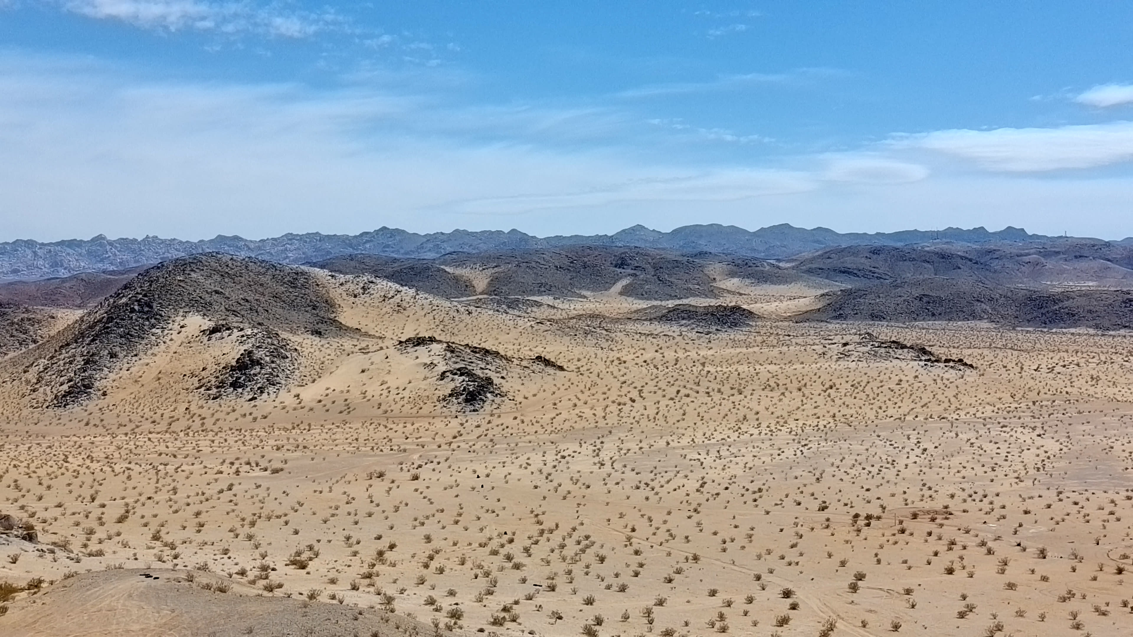A barren desert landscape features rolling hills and sparse vegetation under a clear blue sky.