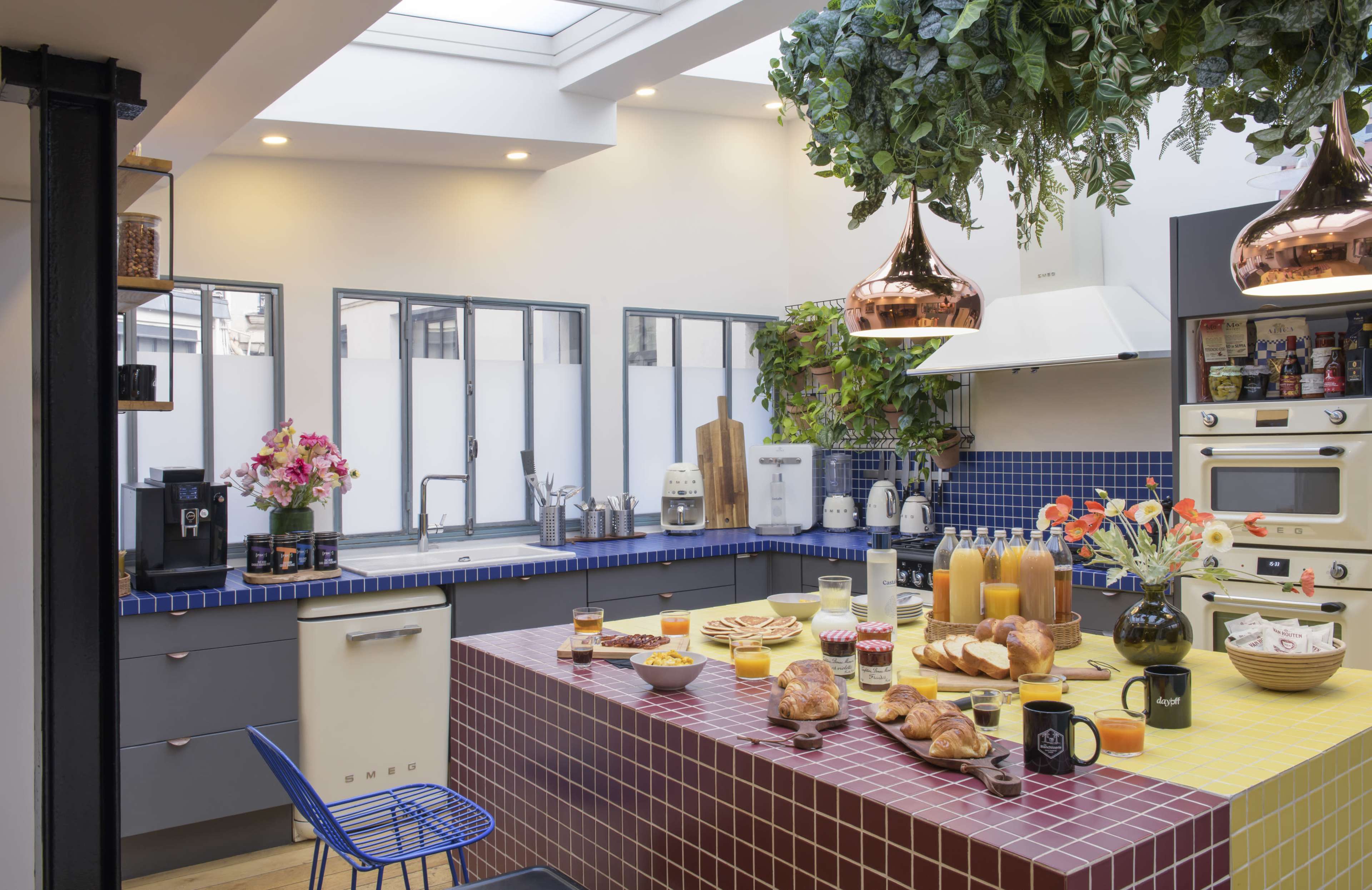 The kitchen features a vibrant mosaic-tiled island with an array of breakfast items, and plants hanging above in a well-lit space with large windows.