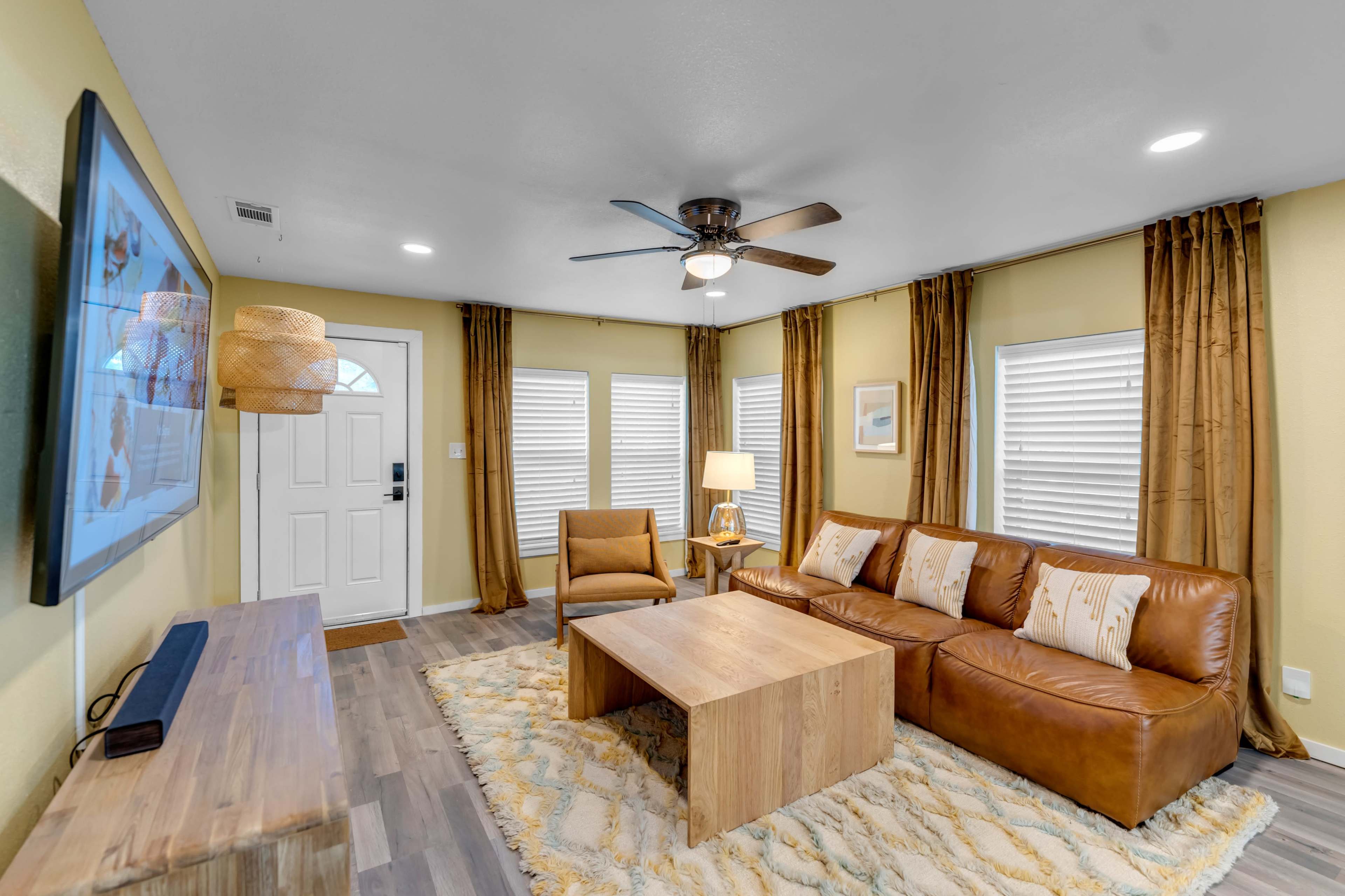 A cozy living room with a leather sofa, a wooden coffee table, and a large TV mounted on the wall, complemented by yellow walls and light-colored window treatments.