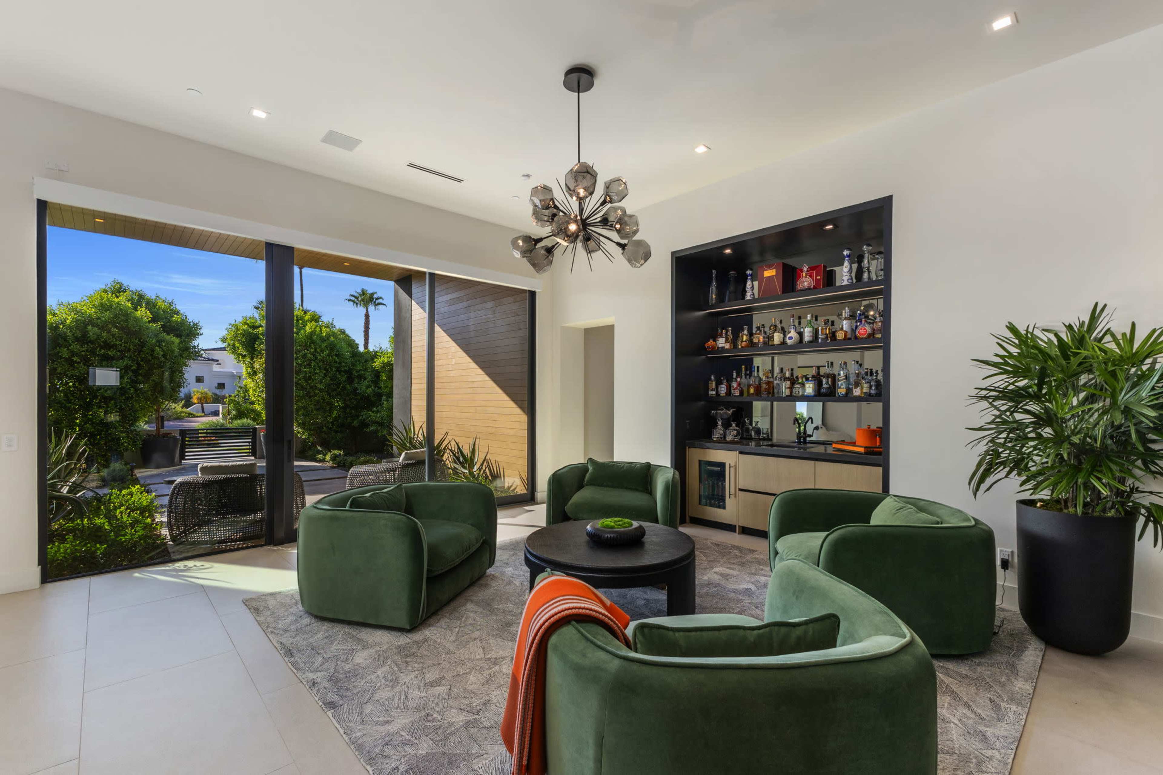 A modern living room features four green armchairs arranged around a circular black table, with a bar shelf and large windows overlooking a garden.