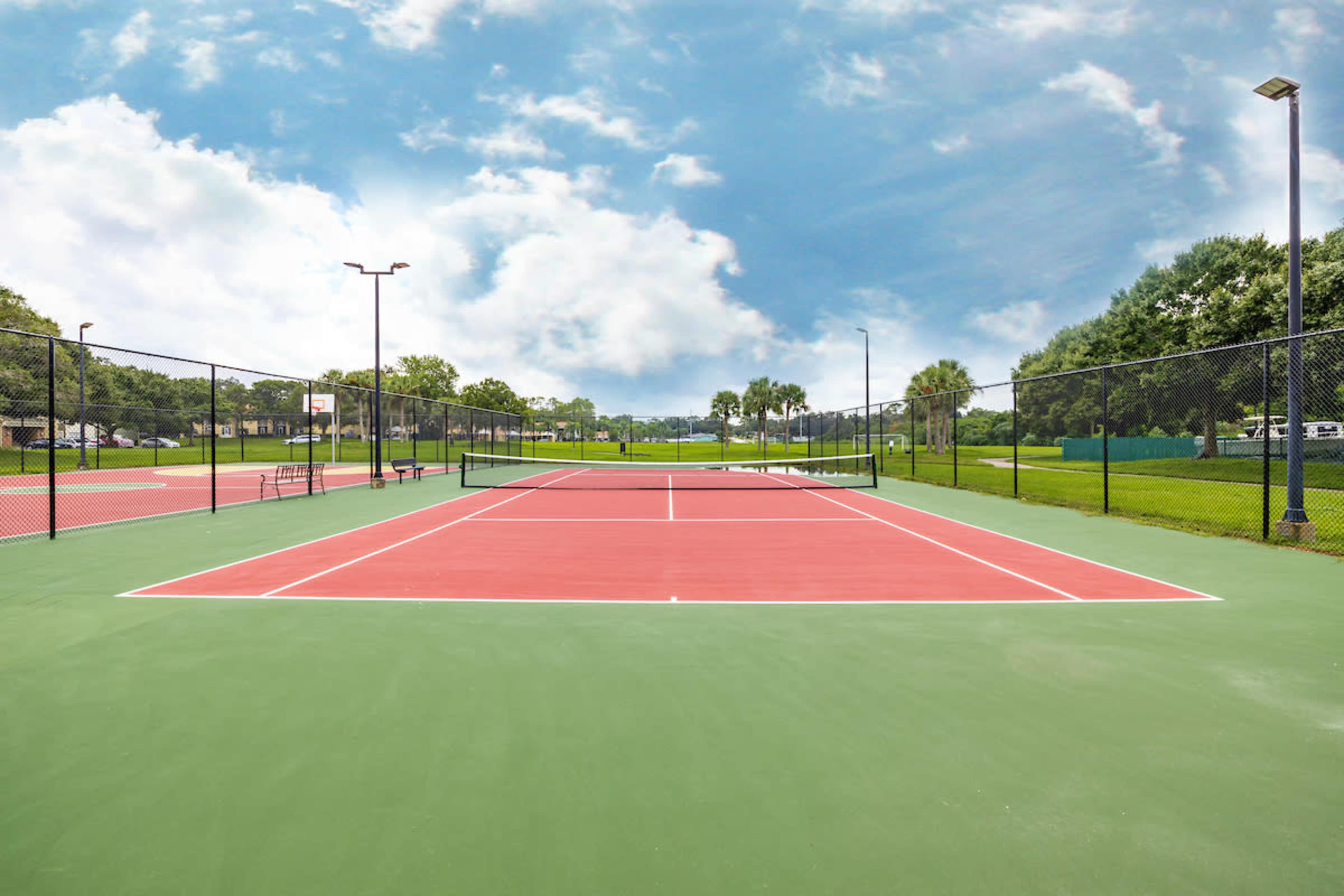 A pair of empty tennis courts surrounded by green trees and under a cloudy blue sky.