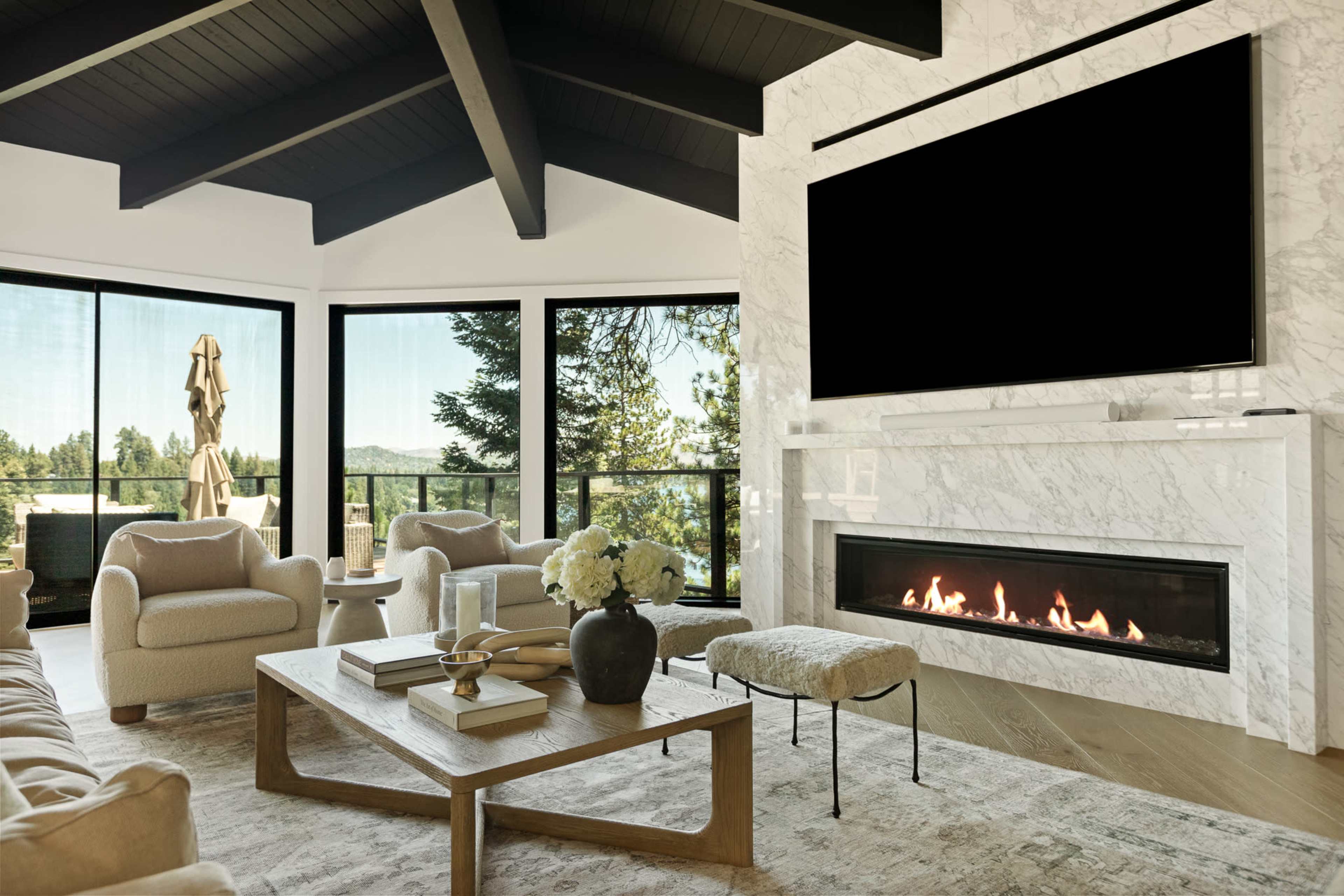 The image shows a modern living room featuring a marble fireplace, large windows, and contemporary furniture with a view of greenery outside.
