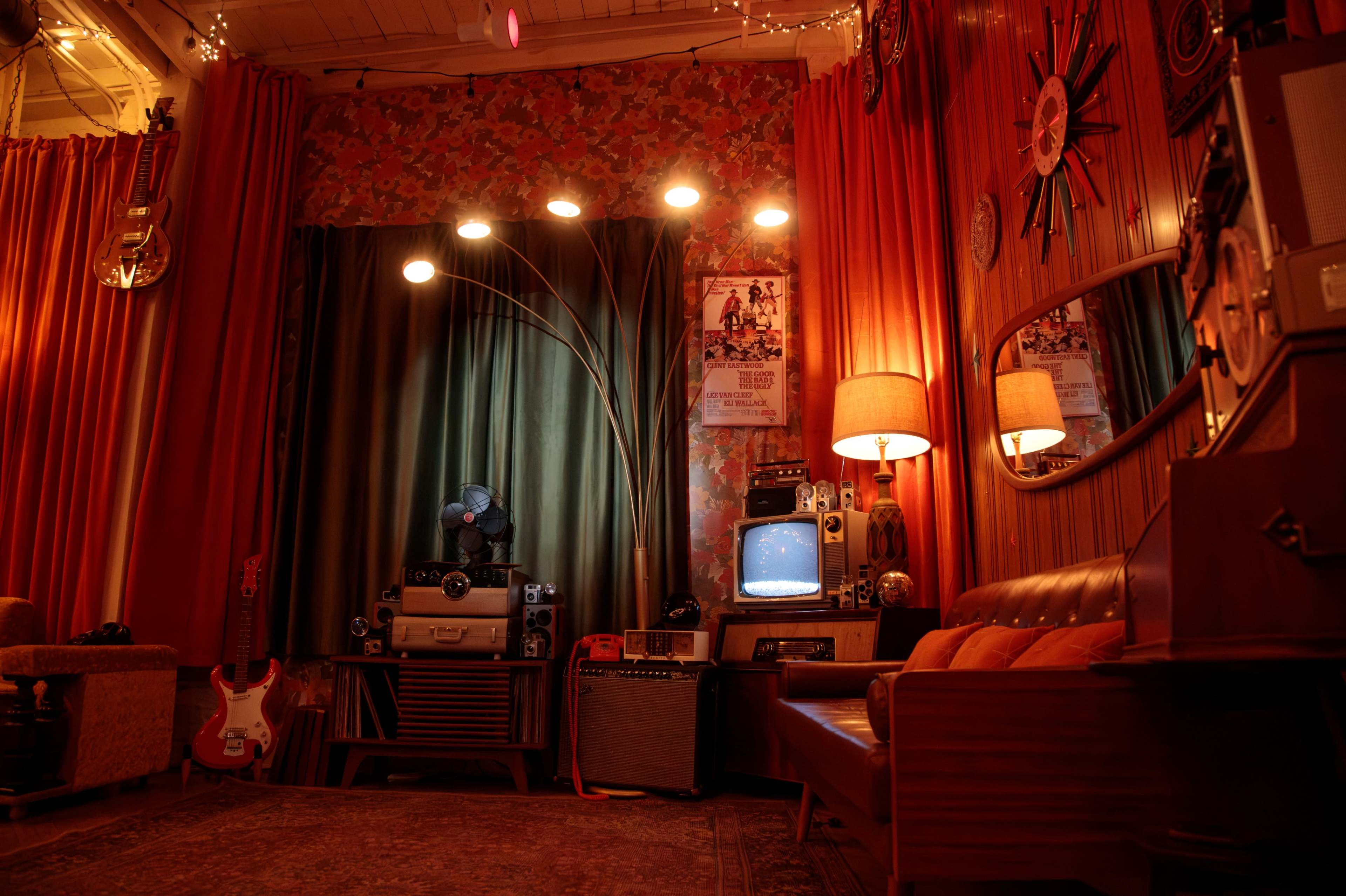 The image shows a retro-style room with a large green curtain, vintage furniture, and various musical instruments and electronics arranged around the space.