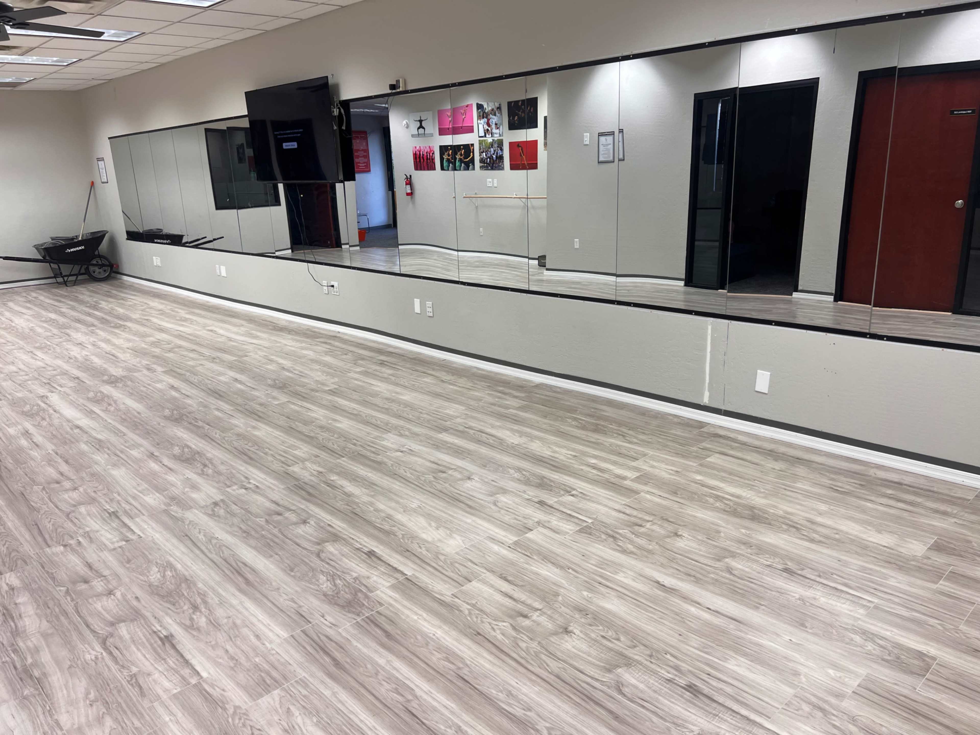 The image shows a spacious, well-lit room with wooden flooring, a long mirror on one wall, and a television mounted nearby.