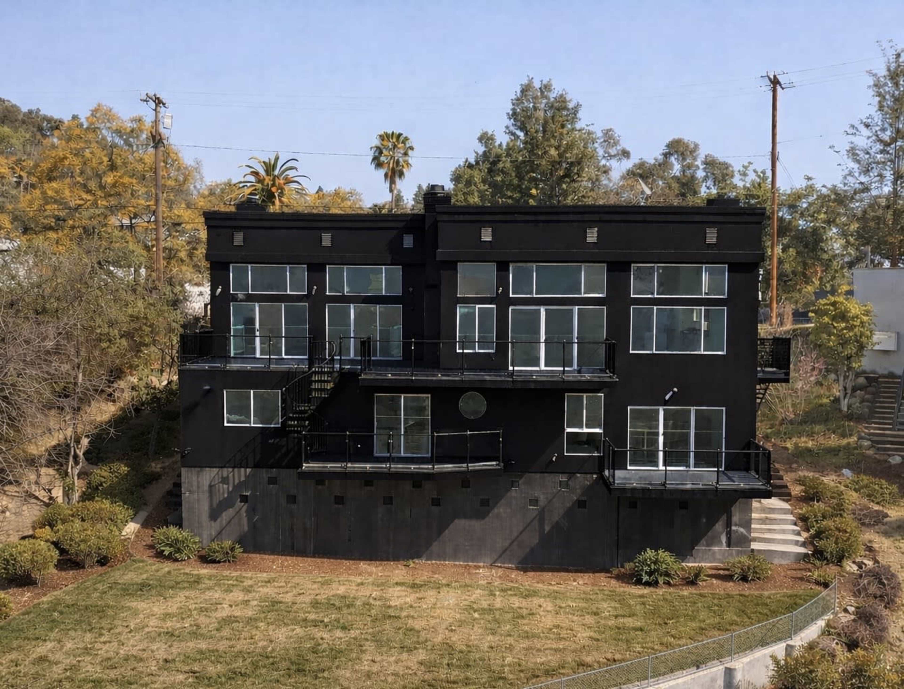 The image shows a large, modern black house with multiple levels and large windows surrounded by greenery.