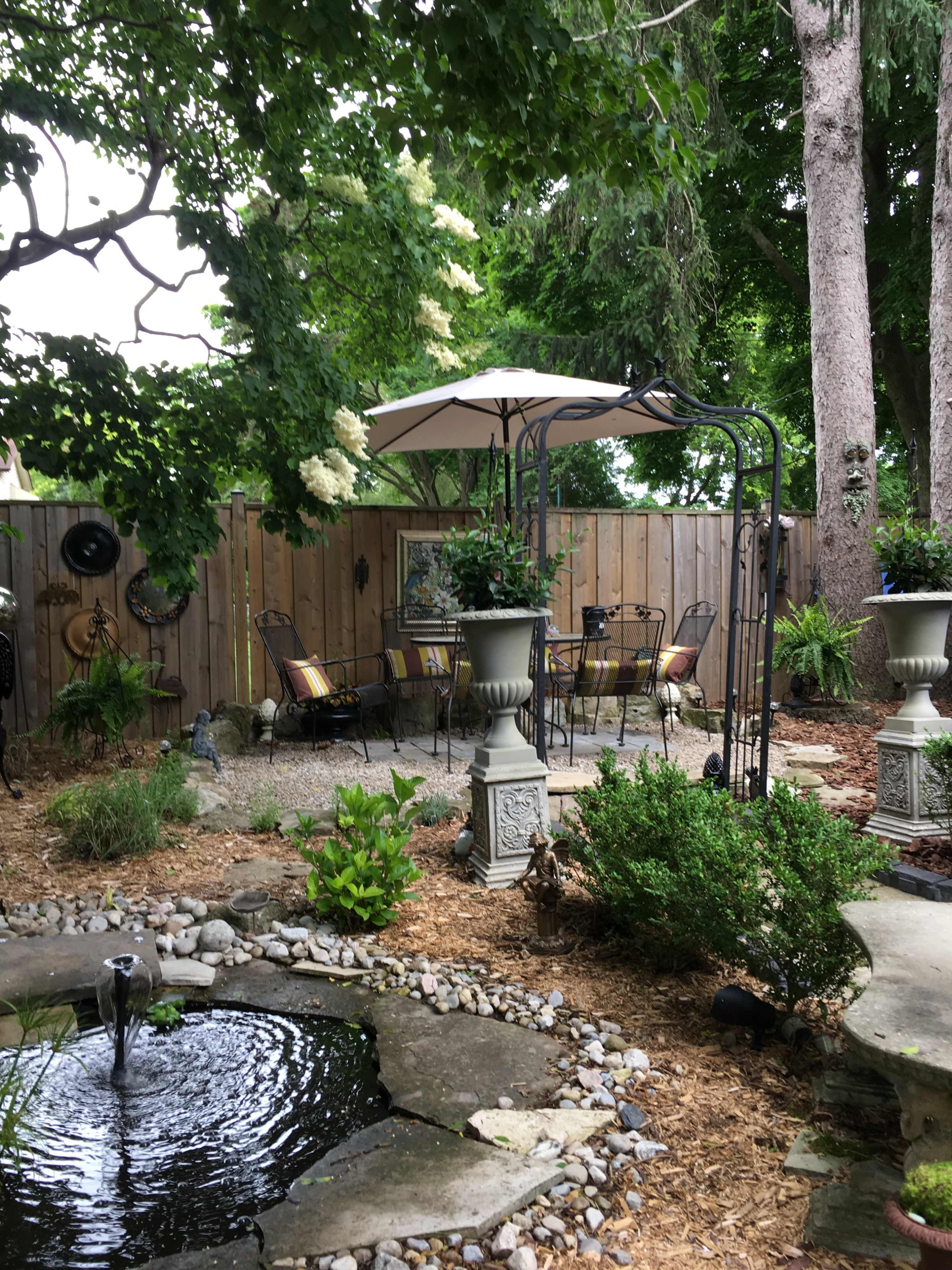 A shaded outdoor seating area features a gazebo, stone fountain, and potted plants amidst gravel and mulch landscaping.