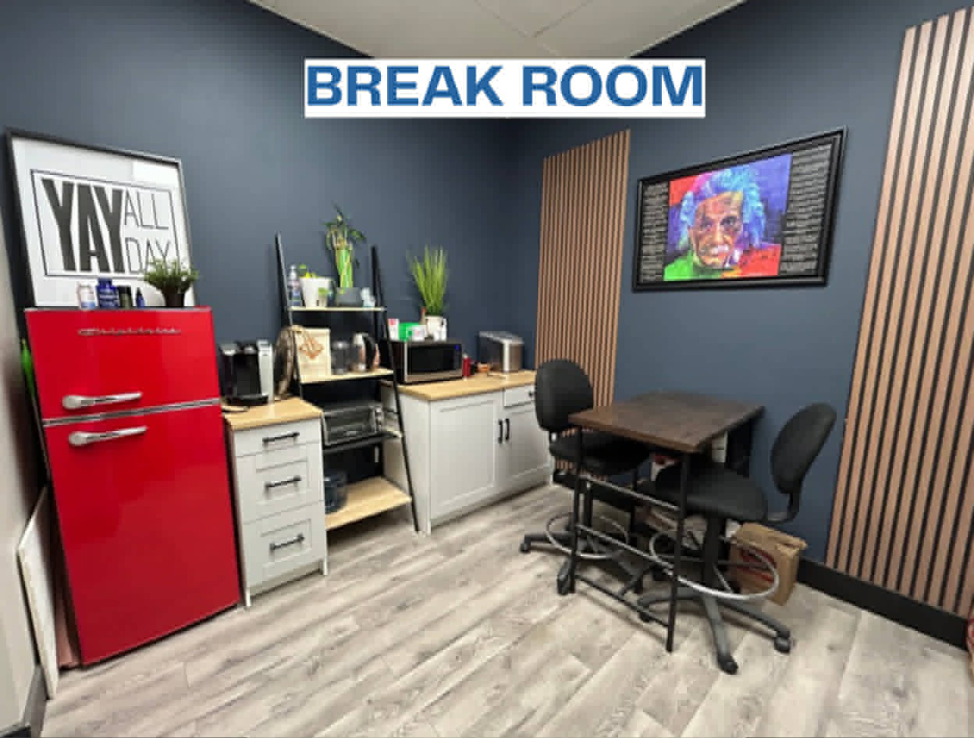 The image shows a break room featuring a red refrigerator, a small kitchen area with a microwave and coffee maker, a table with two chairs, and green plants against a blue wall.