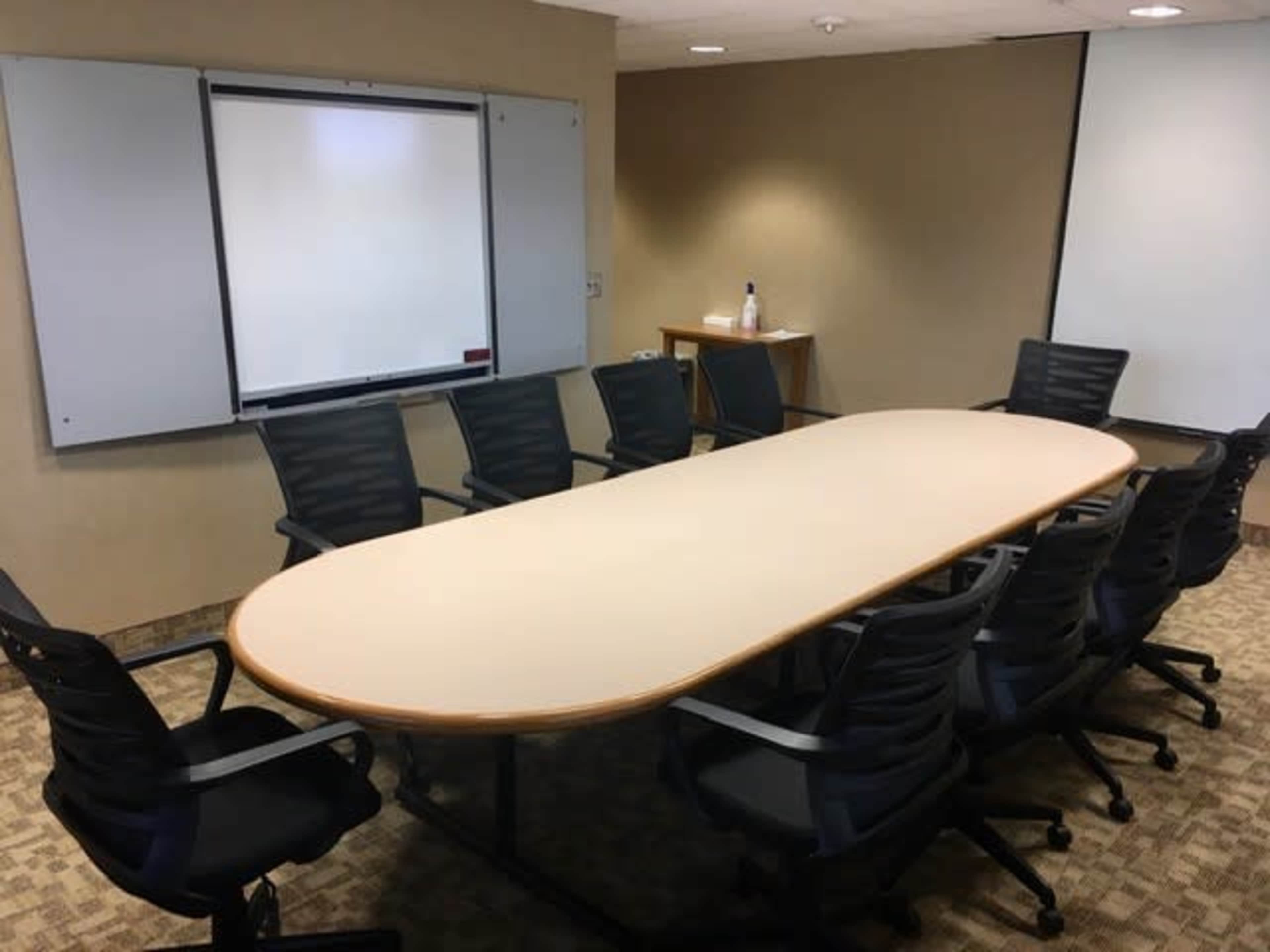 A large, oval table surrounded by eight chairs is situated in a conference room with whiteboards on the walls.