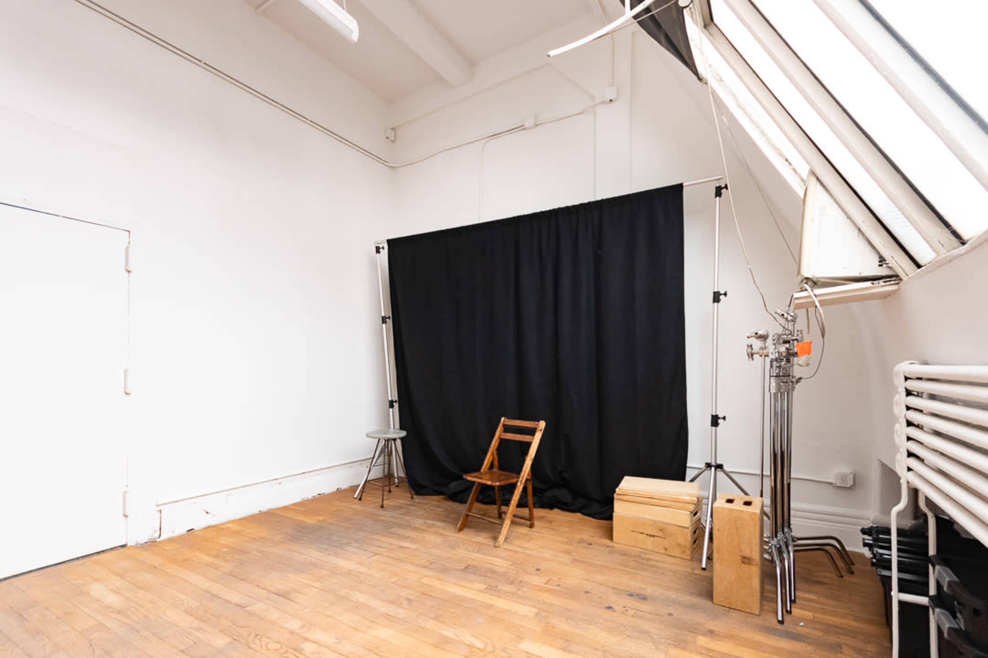 A spacious, well-lit photography studio with a black backdrop, a wooden chair, a stool, and a wooden box on the floor.