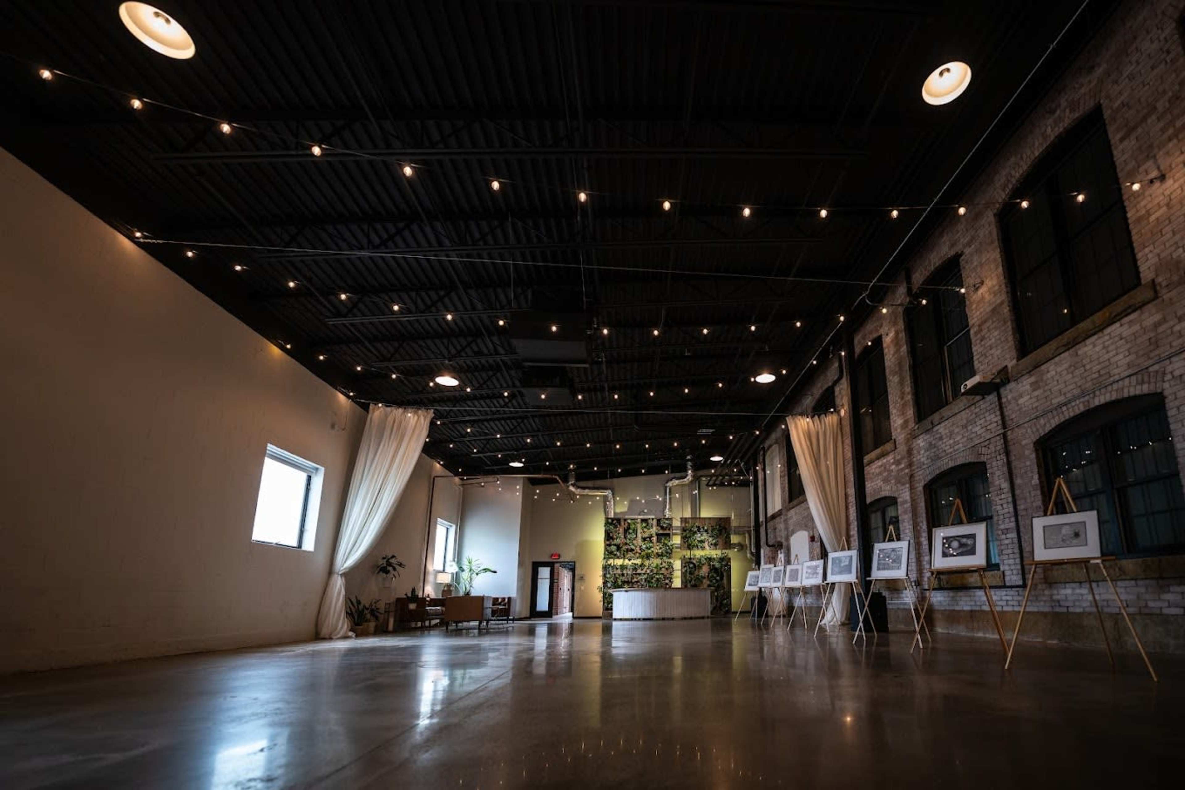 The image shows a spacious, well-lit interior of an art gallery featuring framed artworks, decorative plants, and exposed brick walls.