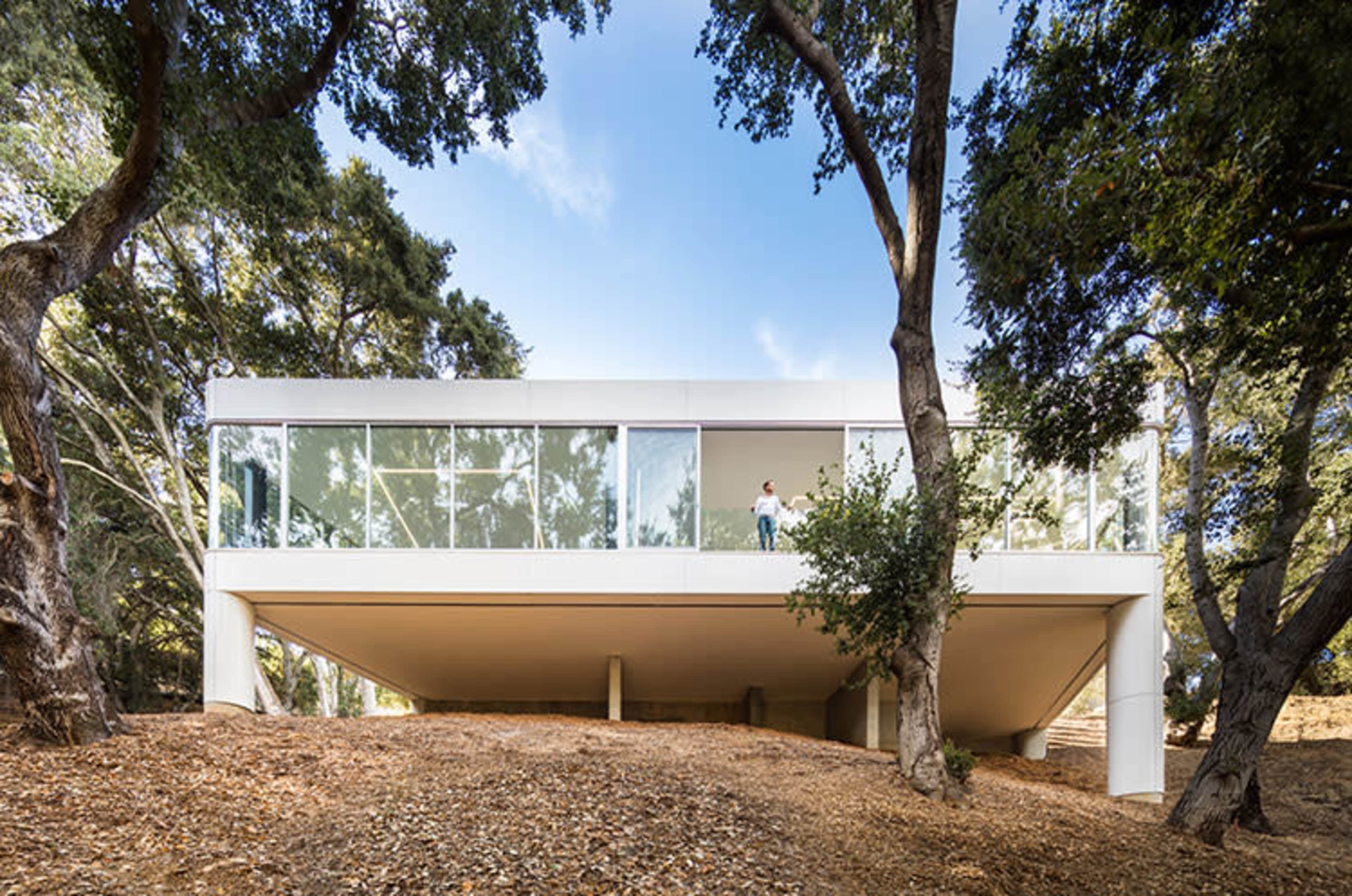 A modern, glass-enclosed house stands elevated on concrete pillars amid trees on a sloping hillside.