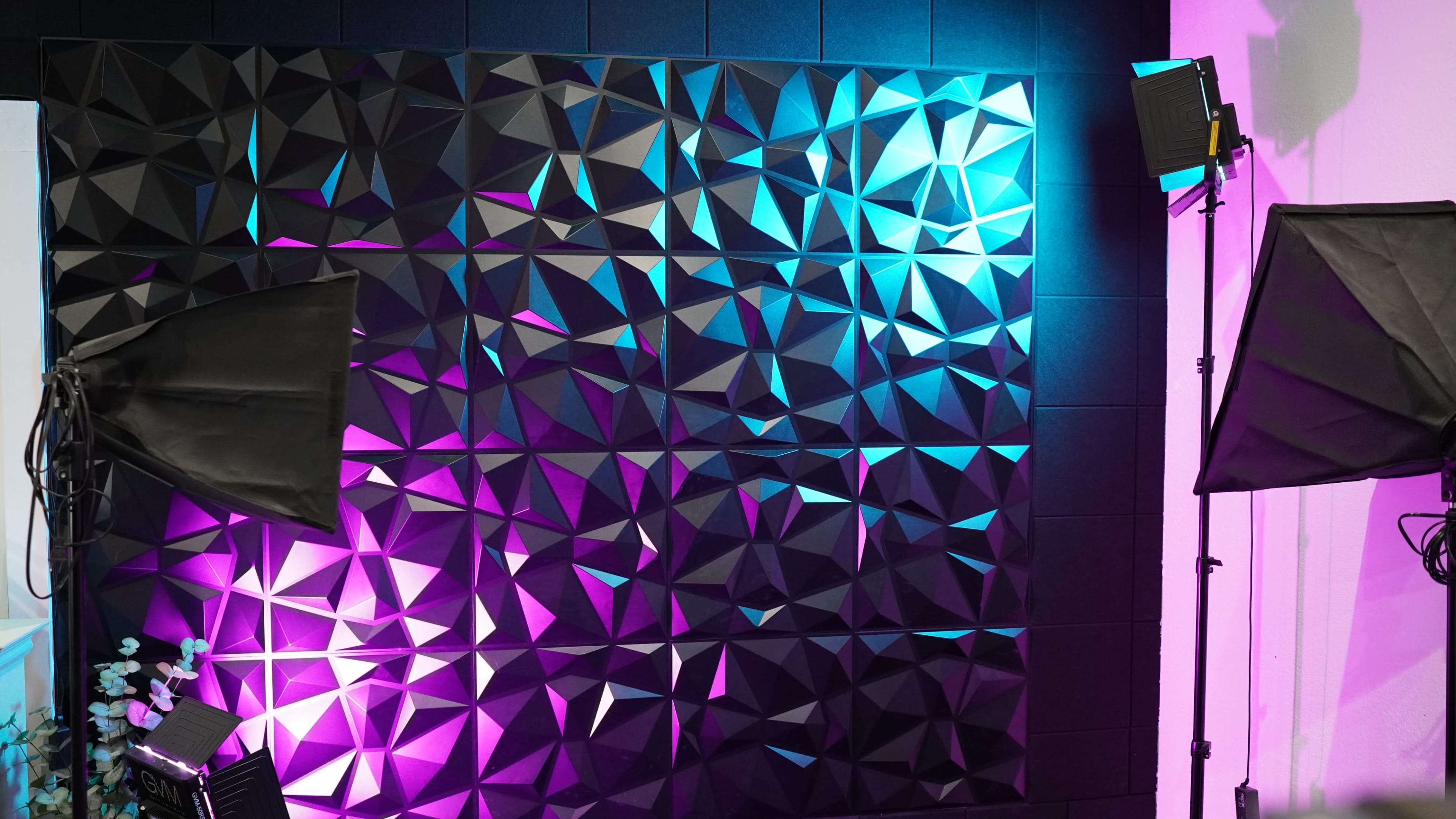 A textured wall with geometric patterns illuminated by blue and purple lights, accompanied by two photography softboxes.