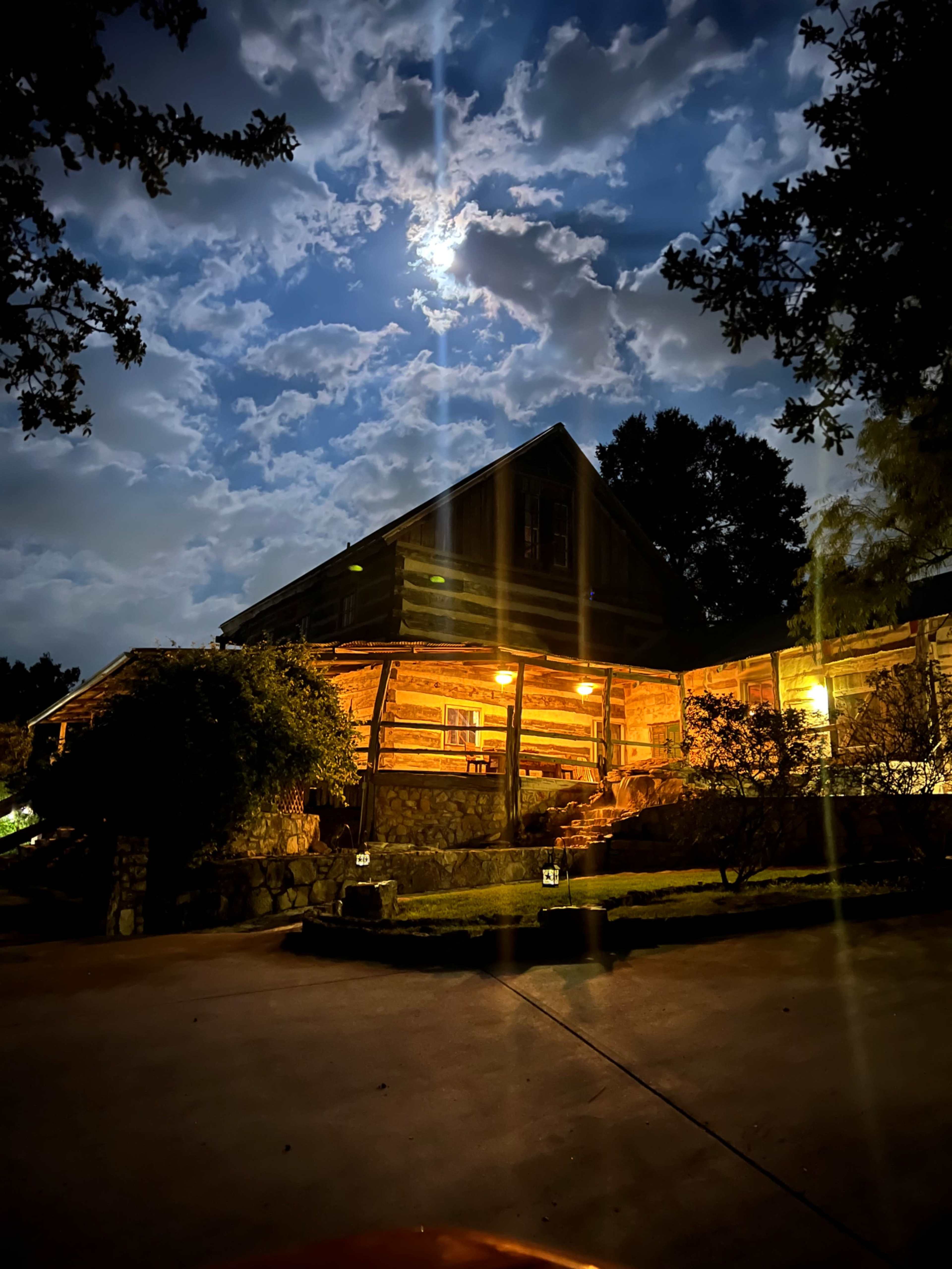 A log cabin with warm lights illuminates the entrance under a cloudy night sky, featuring the moon partially visible among the clouds.