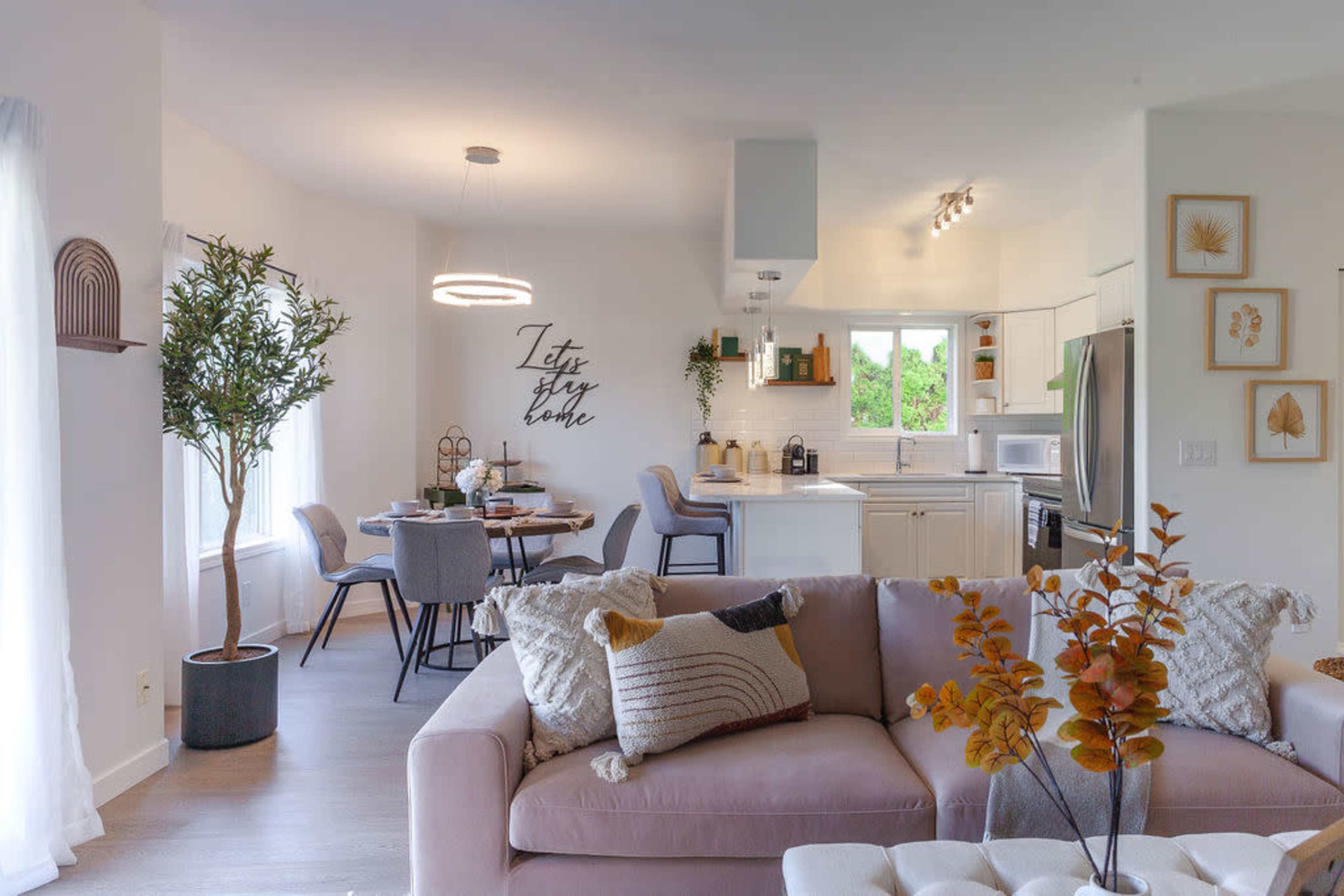 A modern living space with a light-filled open kitchen and dining area, featuring a sectional sofa, a dining table set for four, and decorative wall art.