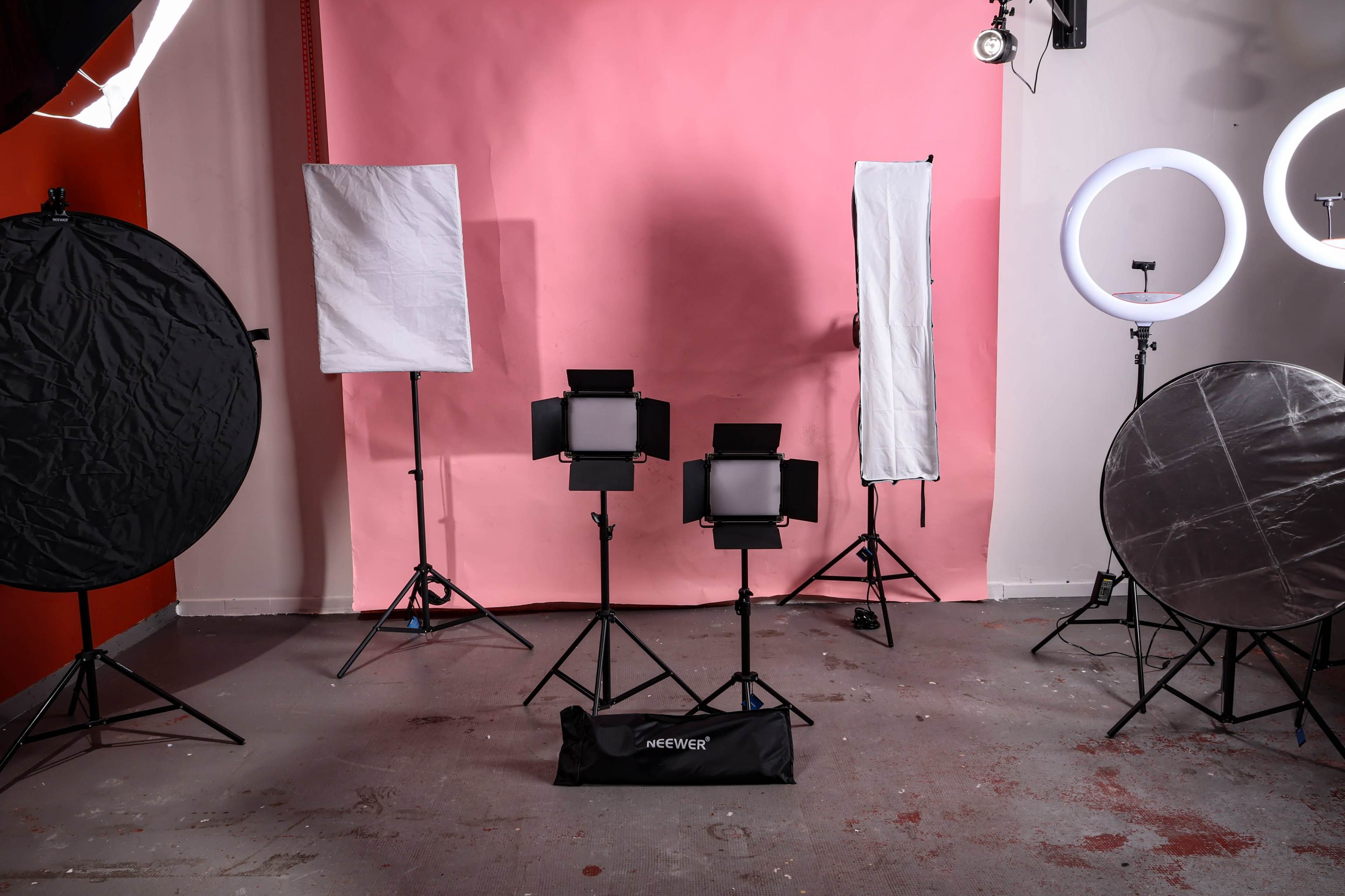 The image shows a photography studio setup with various lighting equipment, including softboxes and LED panels, against a pink backdrop.