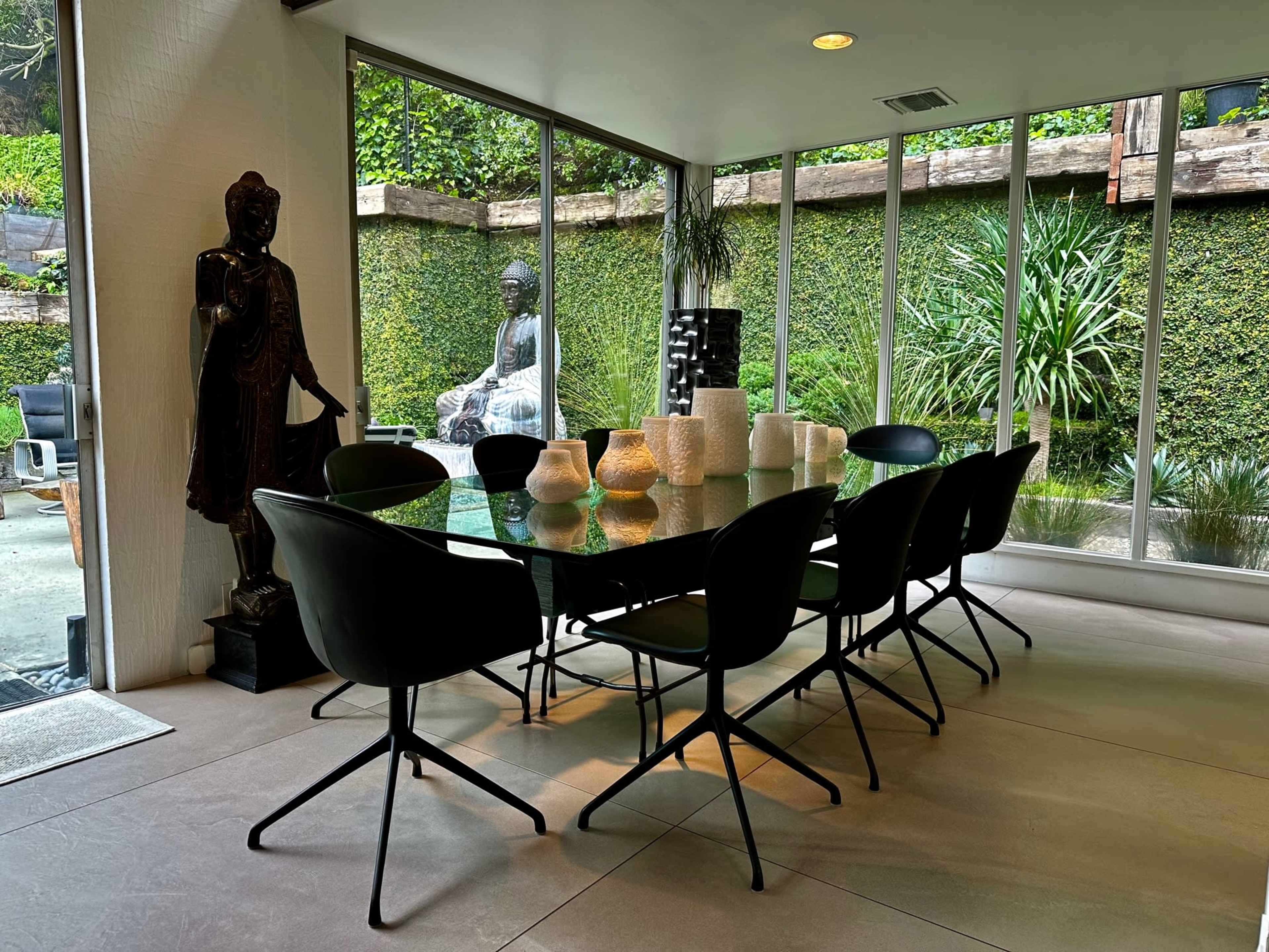 A modern dining room features a glass table surrounded by black chairs, with large windows showcasing a lush green outdoor garden and a statue.