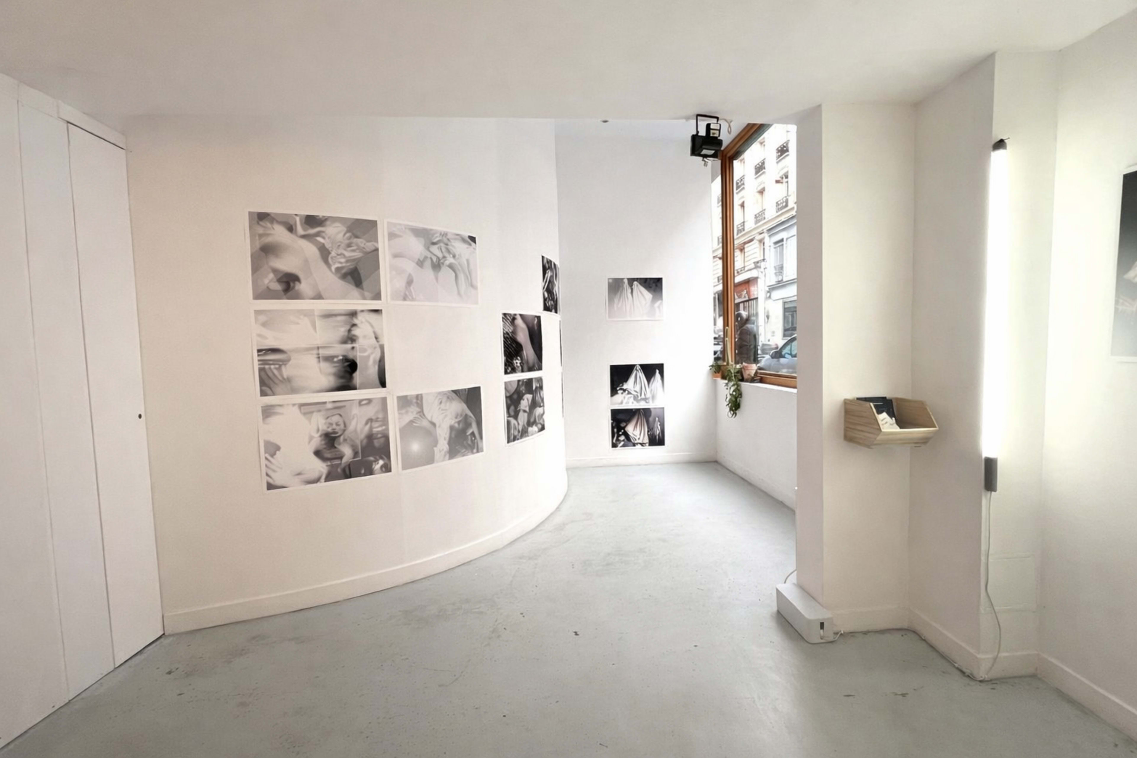 The image shows a white-walled gallery space with several black-and-white photographs displayed on the walls and a small area with natural light coming in from a window.
