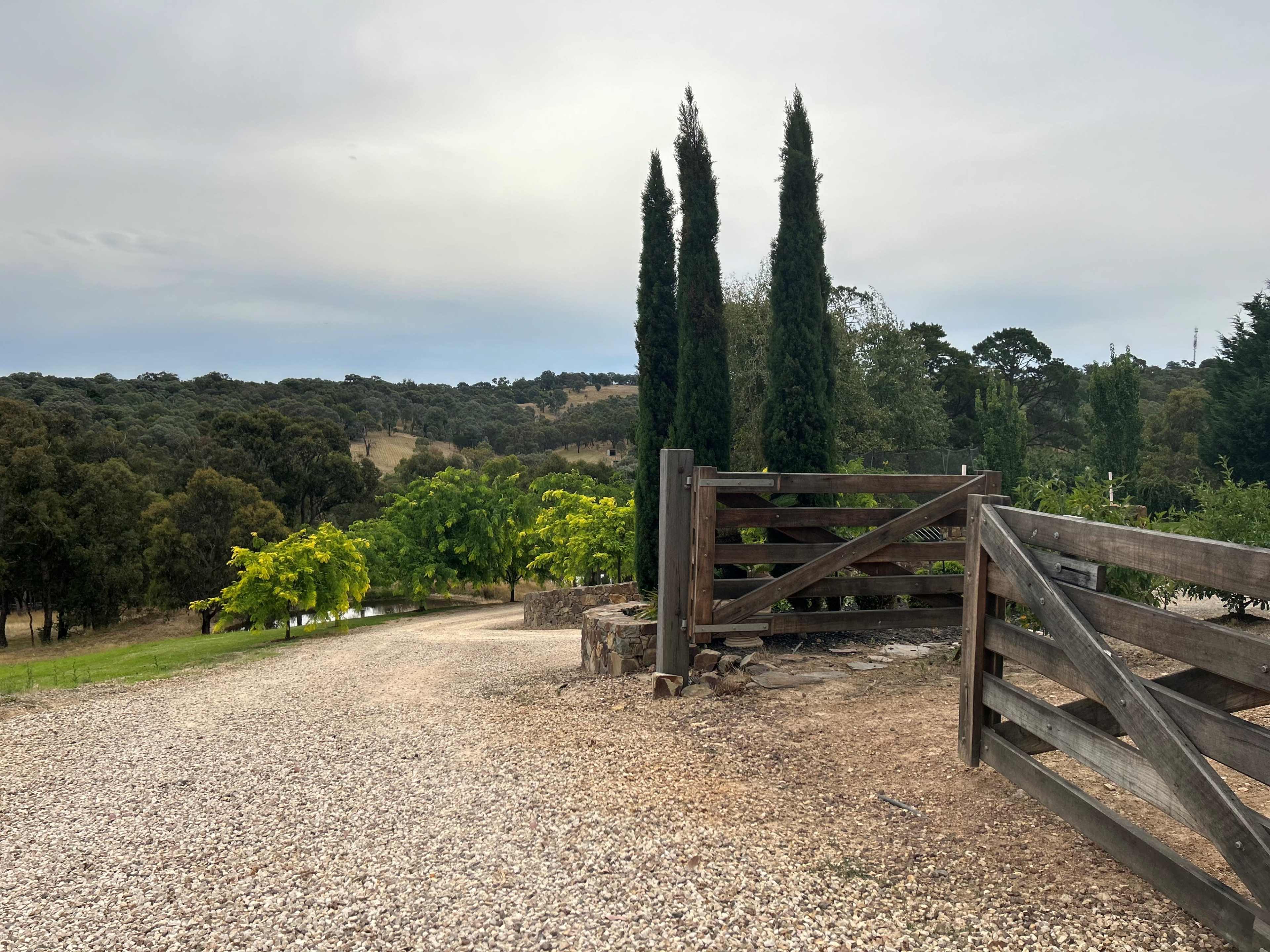 A gravel path leads to a wooden gate surrounded by trees and hills in the background.