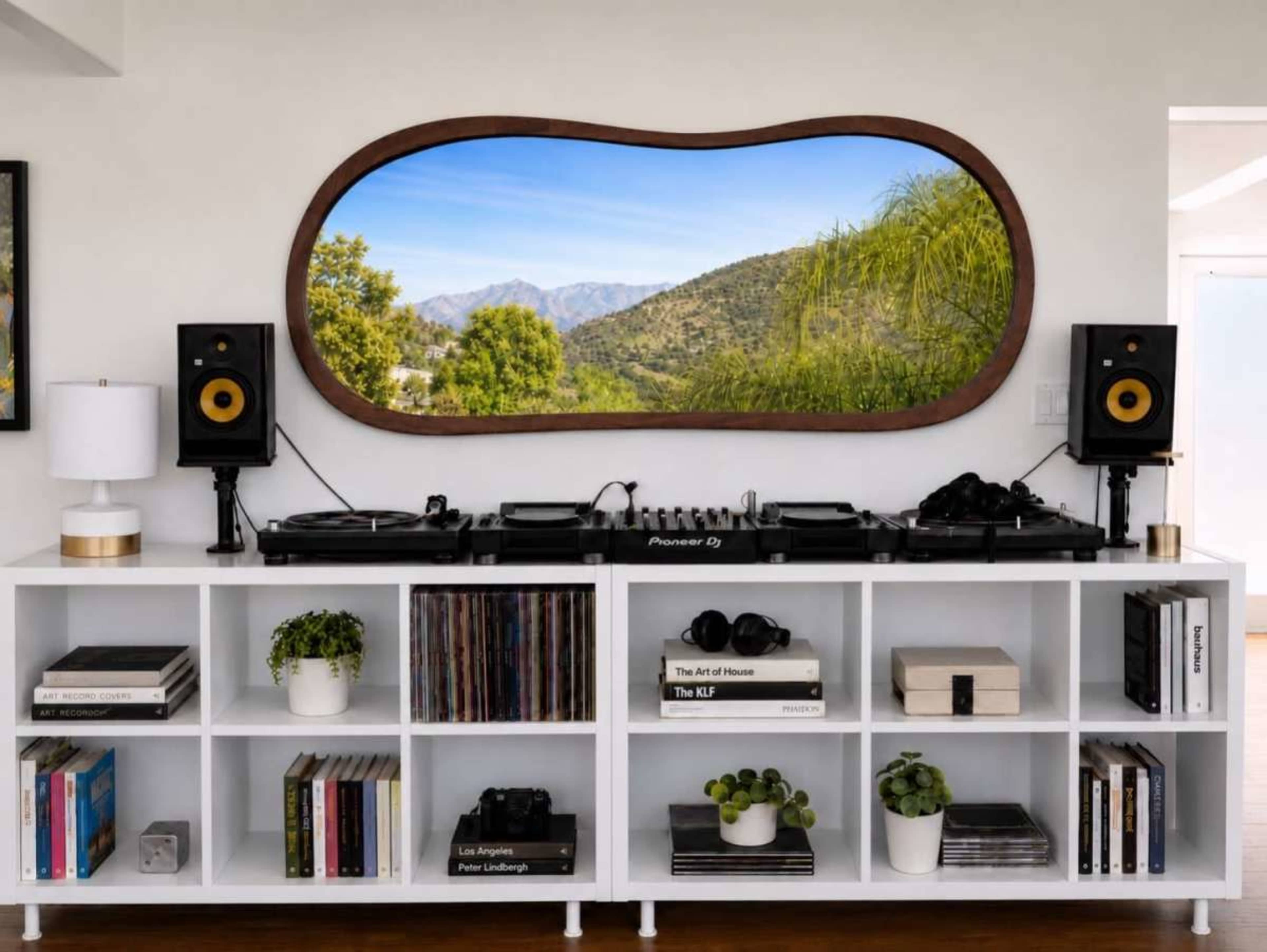 A DJ setup with vinyl records sits on a white shelf, featuring speakers on either side and a large, curved mirror reflecting a mountainous landscape.