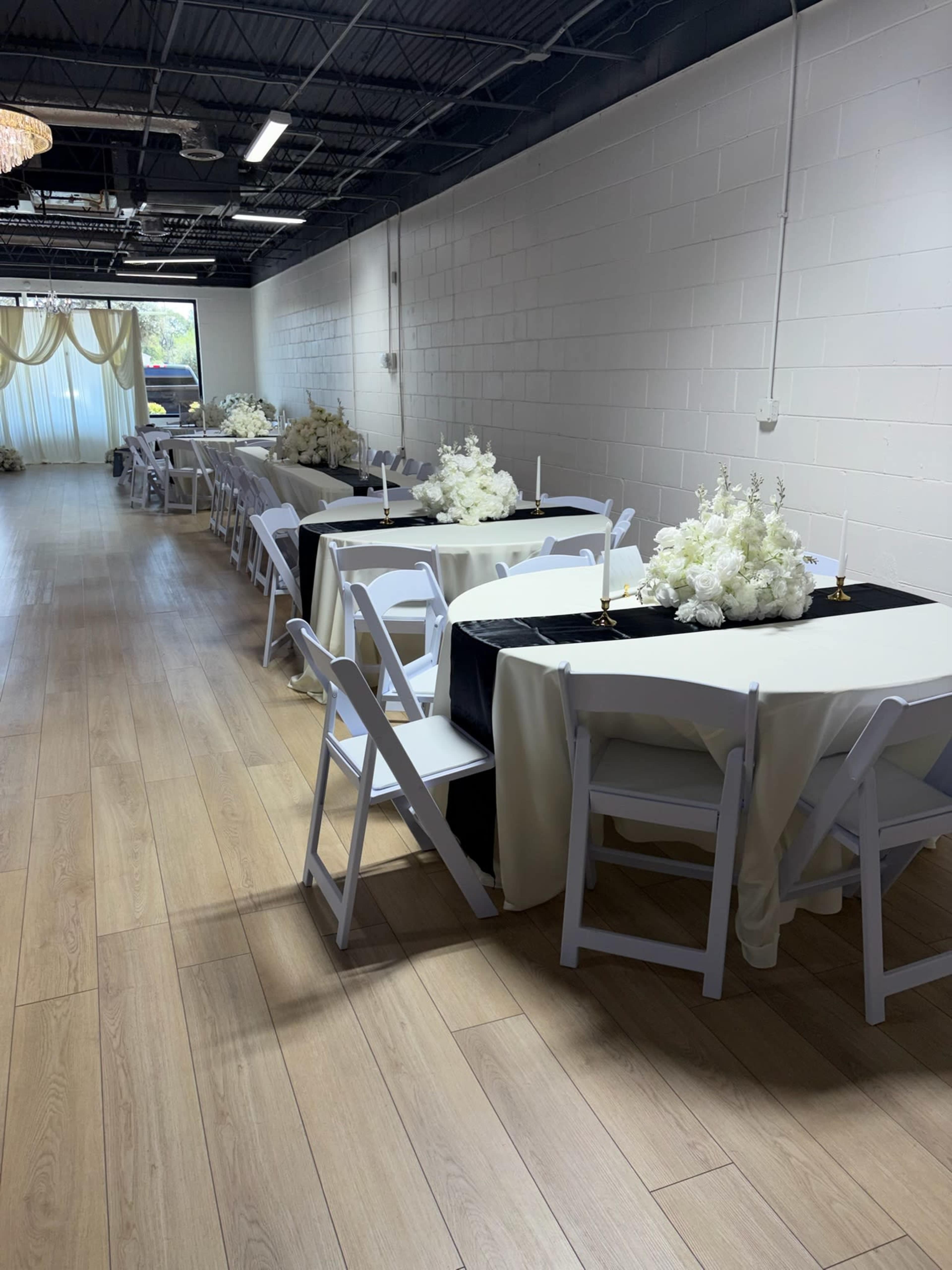 The image shows a long, narrow event space with several rows of covered tables, each adorned with floral arrangements and lit candles, under soft lighting fixtures.