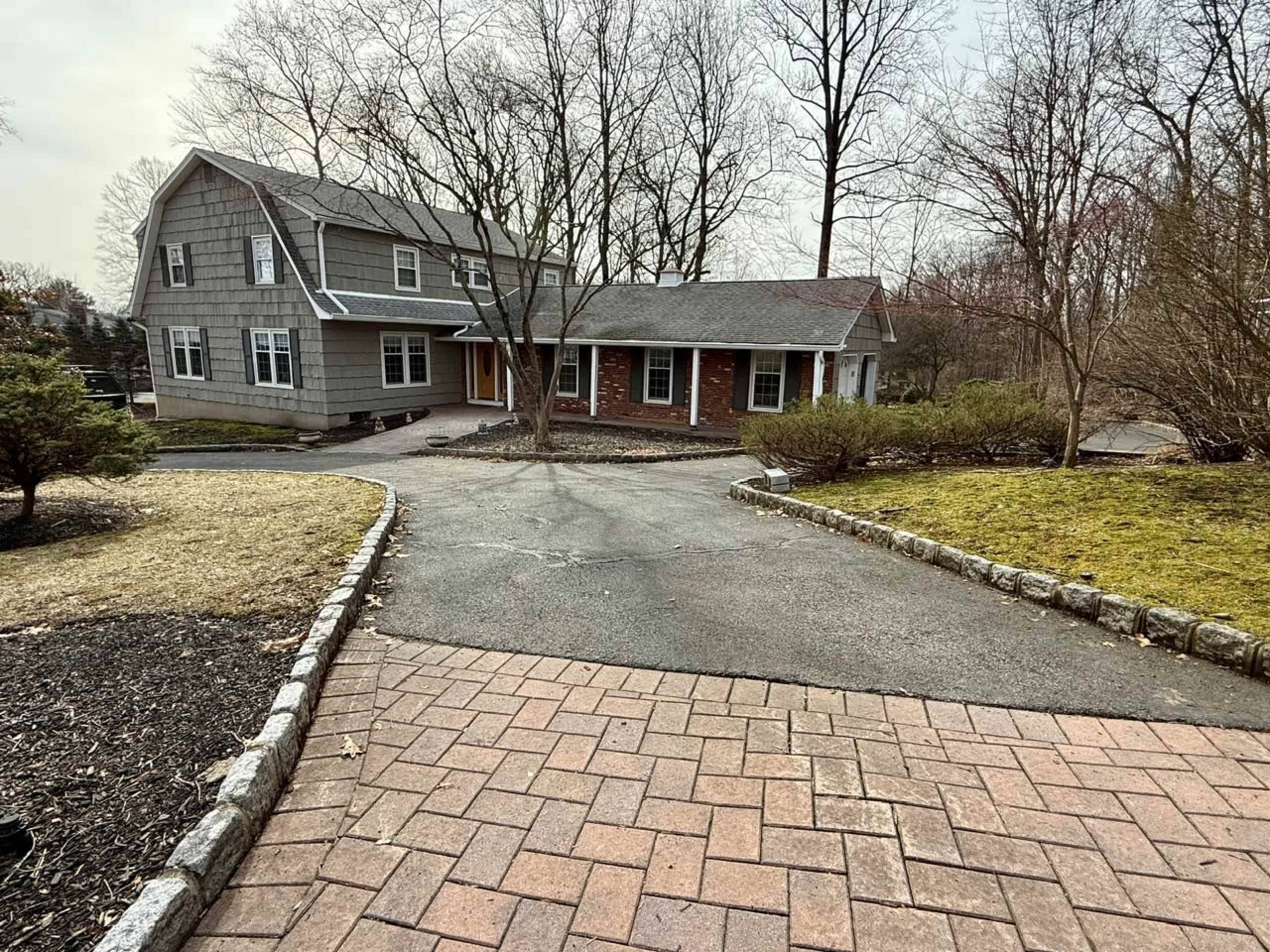 A gray two-story house with a front porch is situated at the end of a paved driveway, surrounded by trees and landscaped bushes.