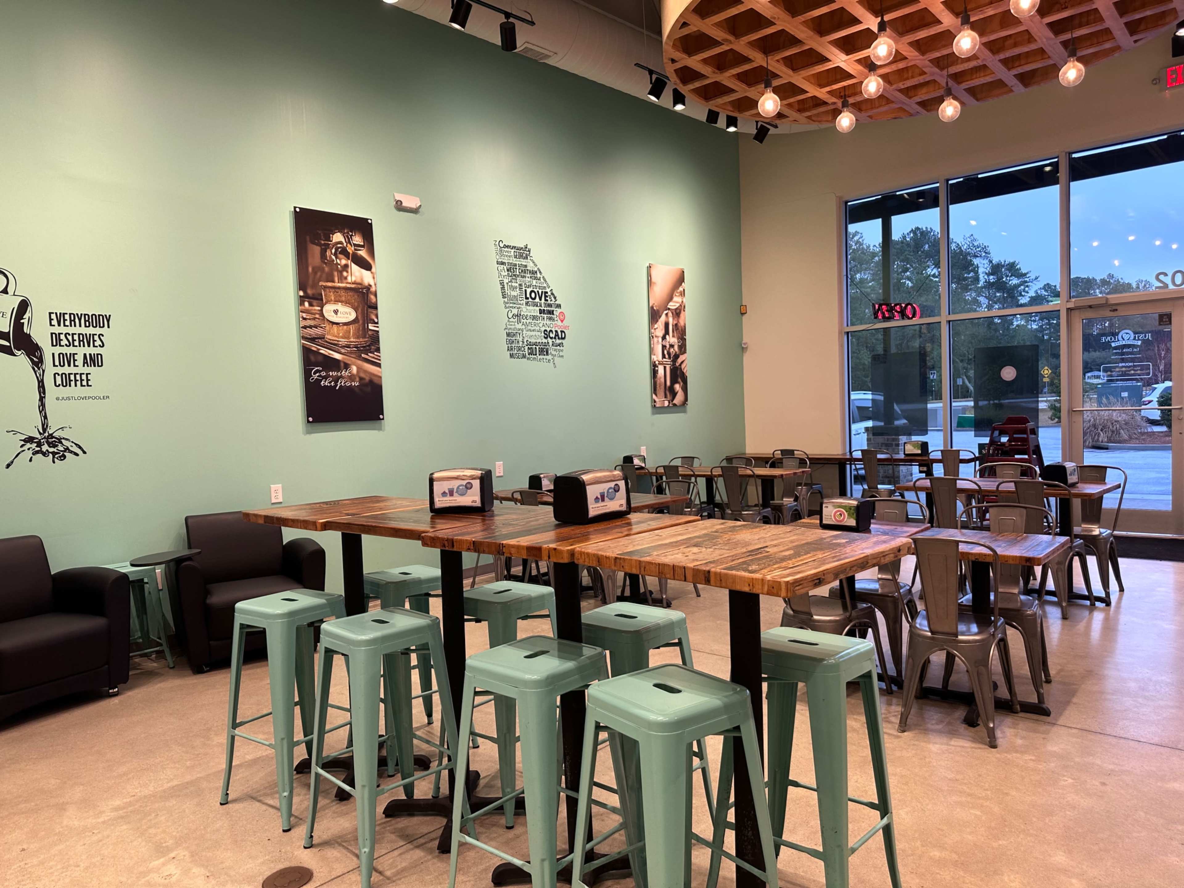 A café interior with a combination of wooden and metal furniture, featuring turquoise stools and rustic tables, along with wall art and large windows.