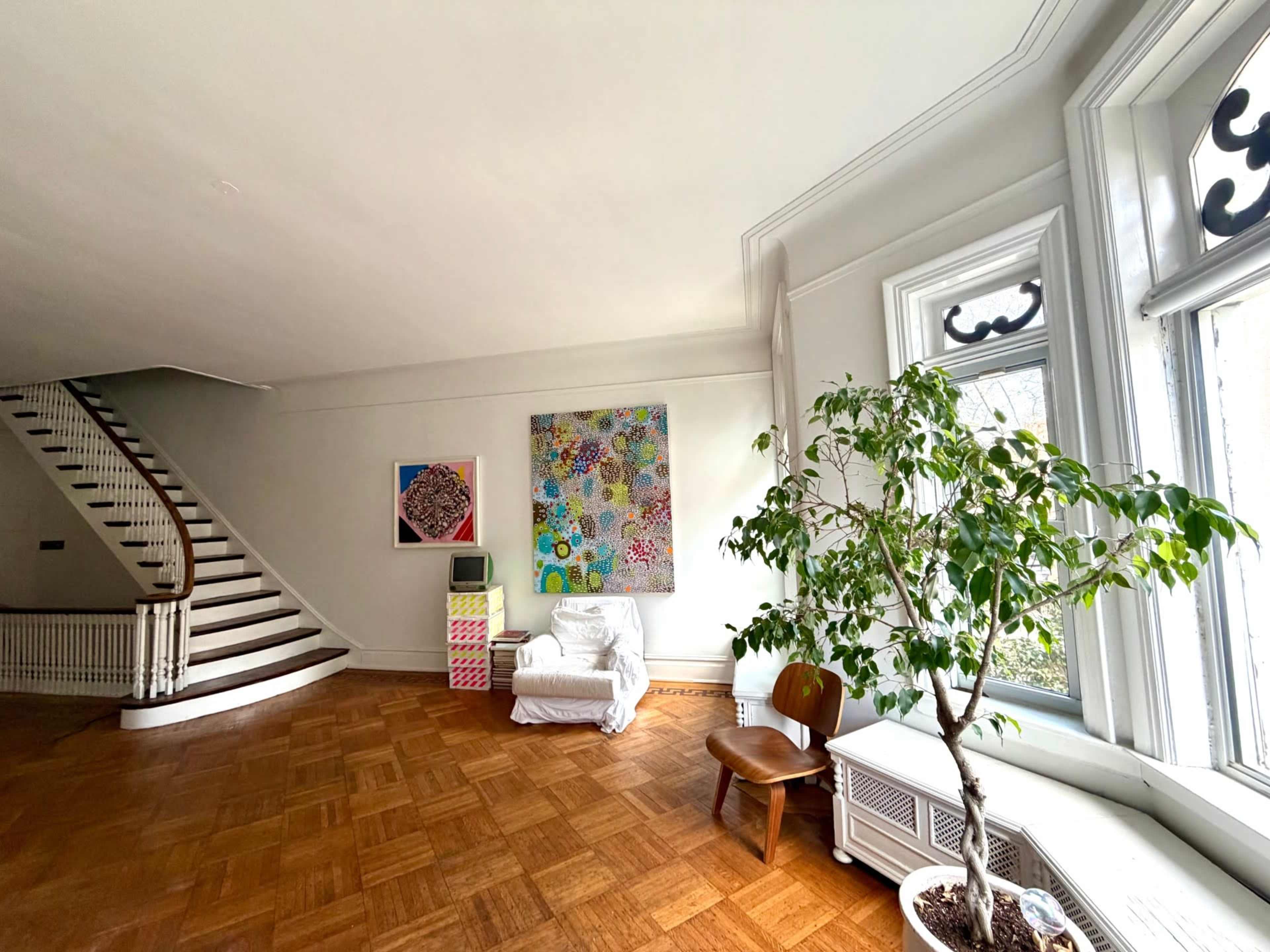 The image shows a spacious room with a wooden floor, a large staircase on the left, a colorful abstract painting on the wall, a white armchair, a potted plant, and a wooden chair near a window.