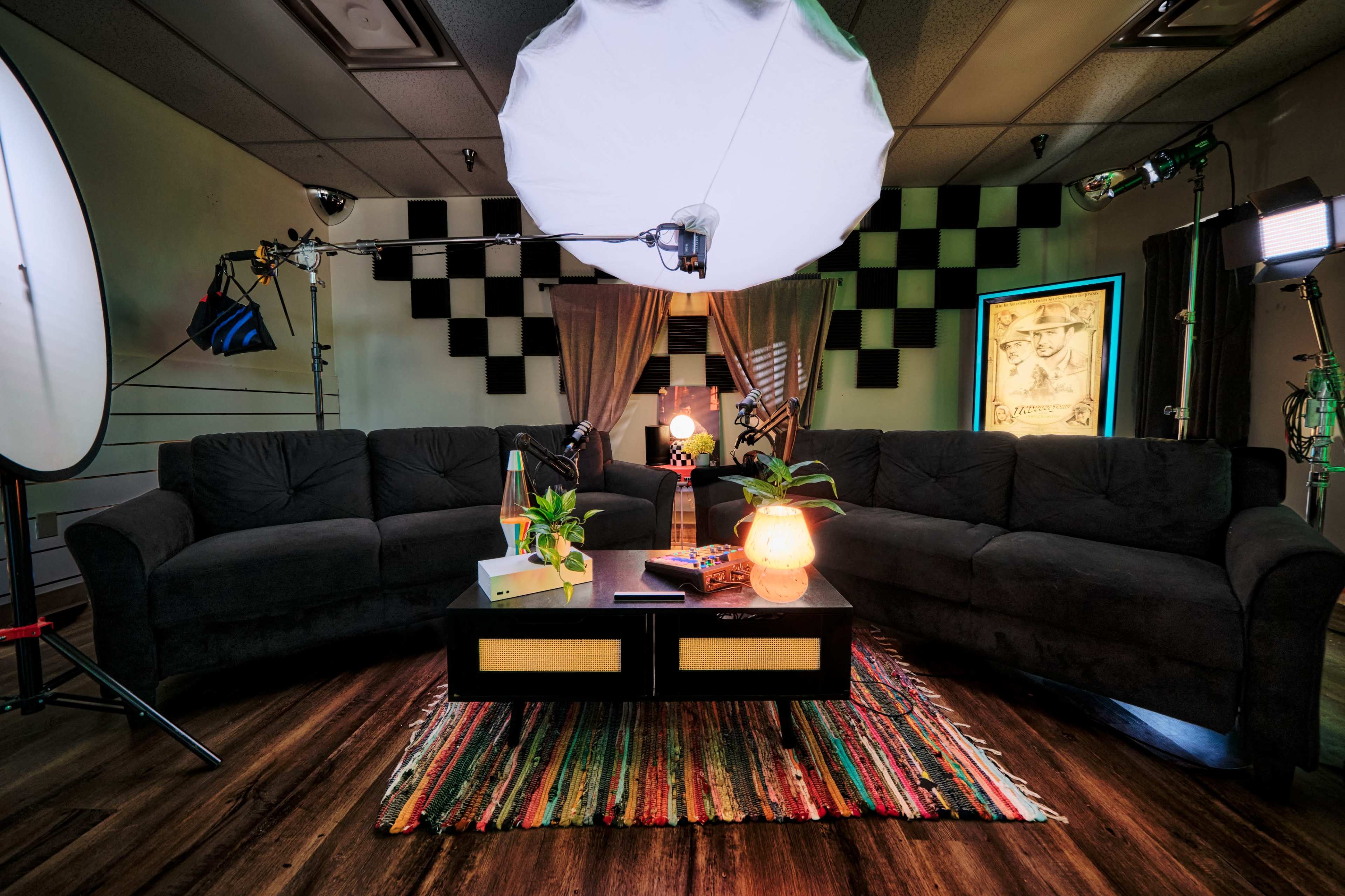 A studio space with two dark sofas arranged around a coffee table, surrounded by various lighting equipment and a colorful rug.