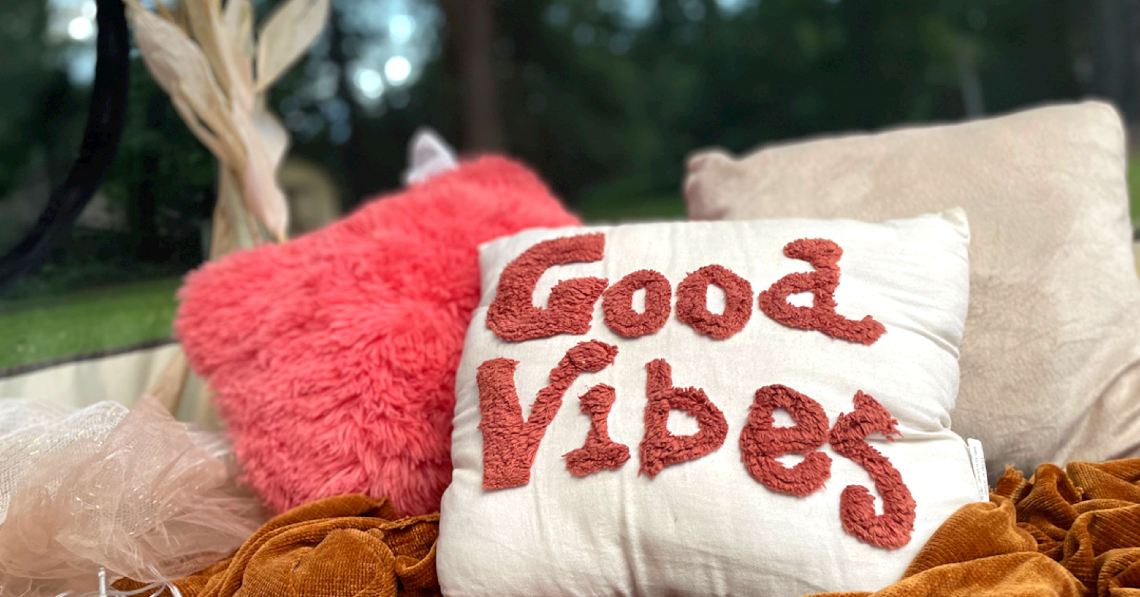 A decorative pillow with the words "Good Vibes" is placed among soft cushions and fabric in a cozy interior setting.