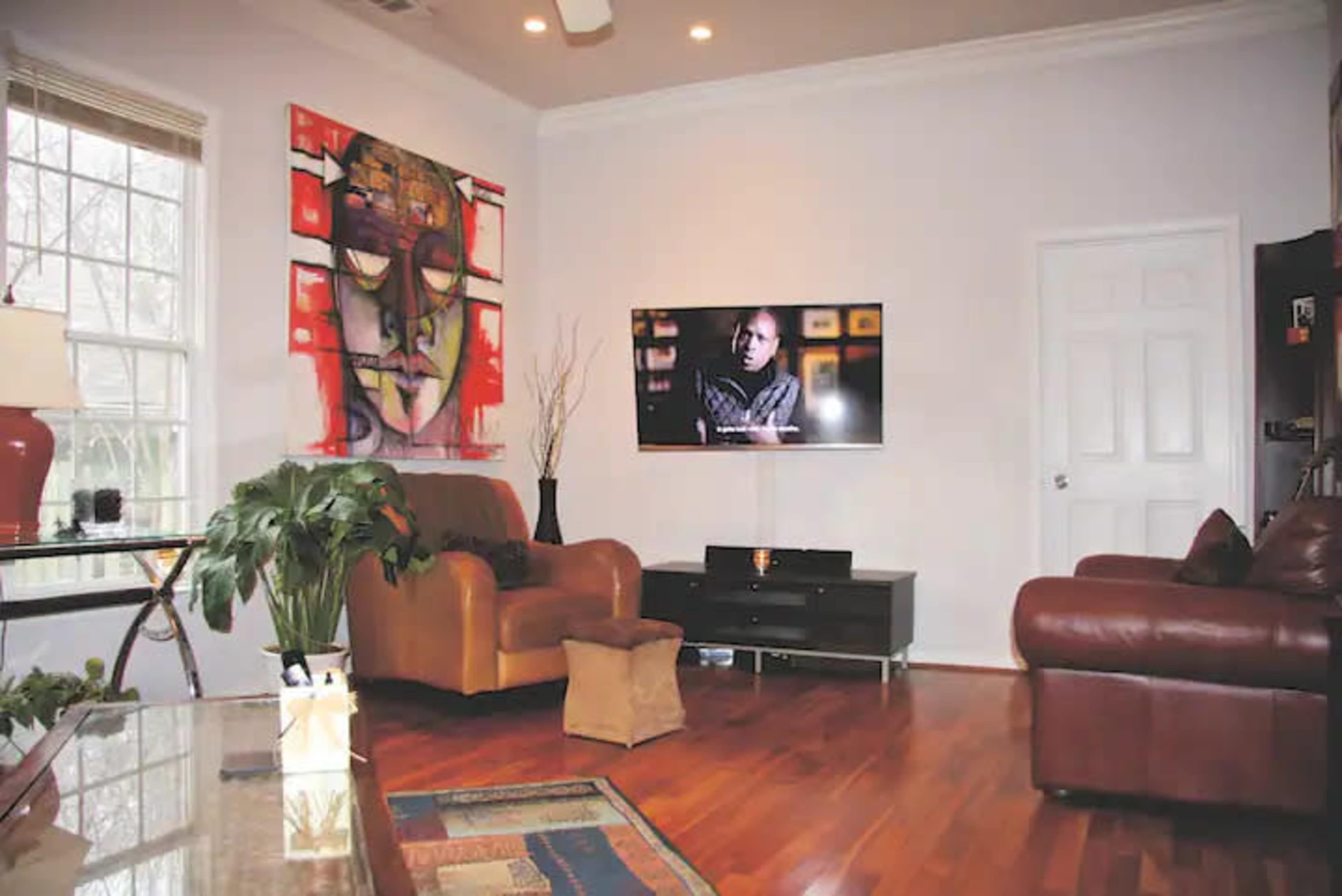 The living room features a large television mounted on the wall across from a brown leather couch and a chair, with a colorful abstract painting and a potted plant in the corner.