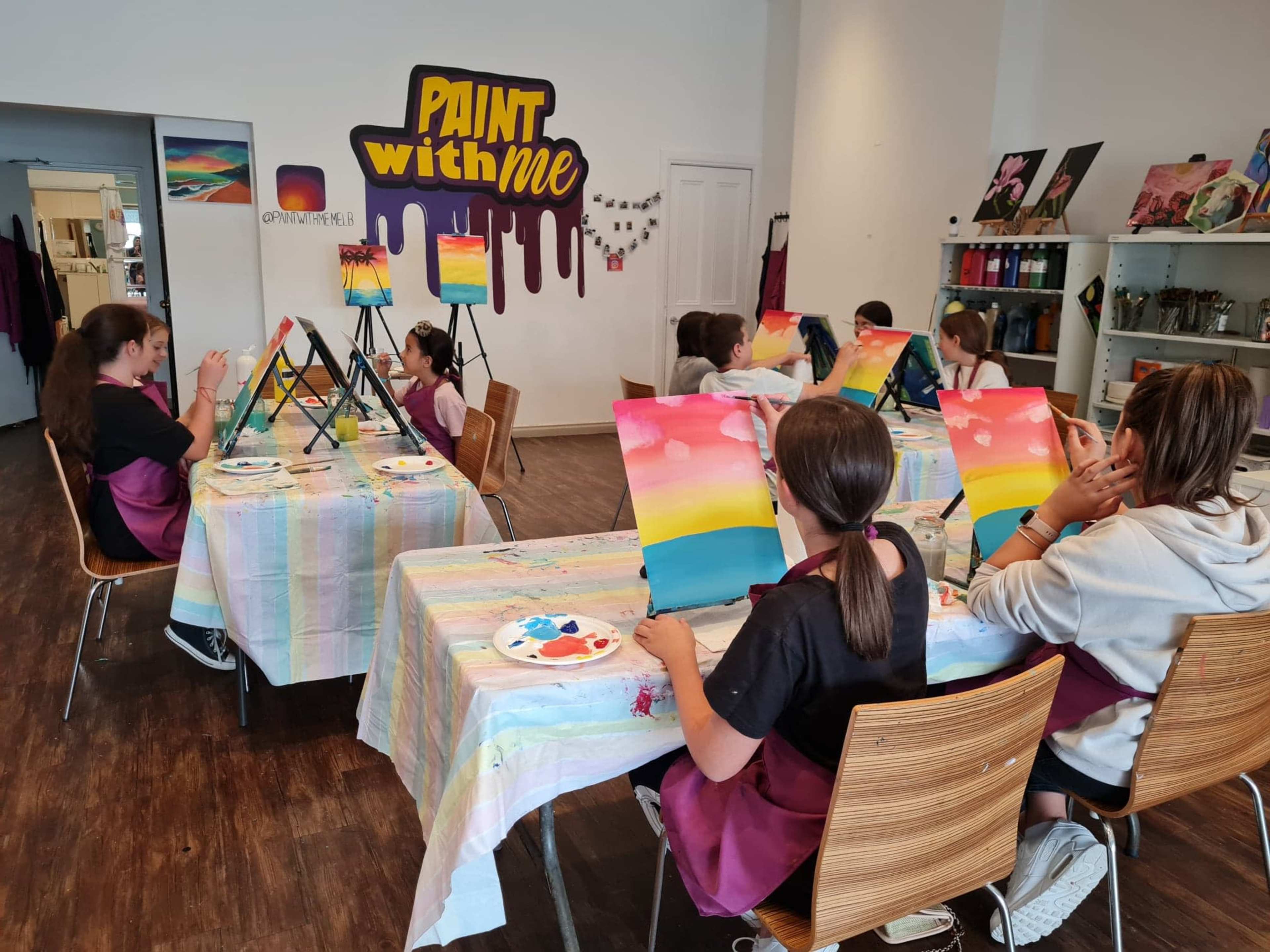 A group of students in aprons are painting on canvases at a classroom with colorful artwork displayed on the walls.