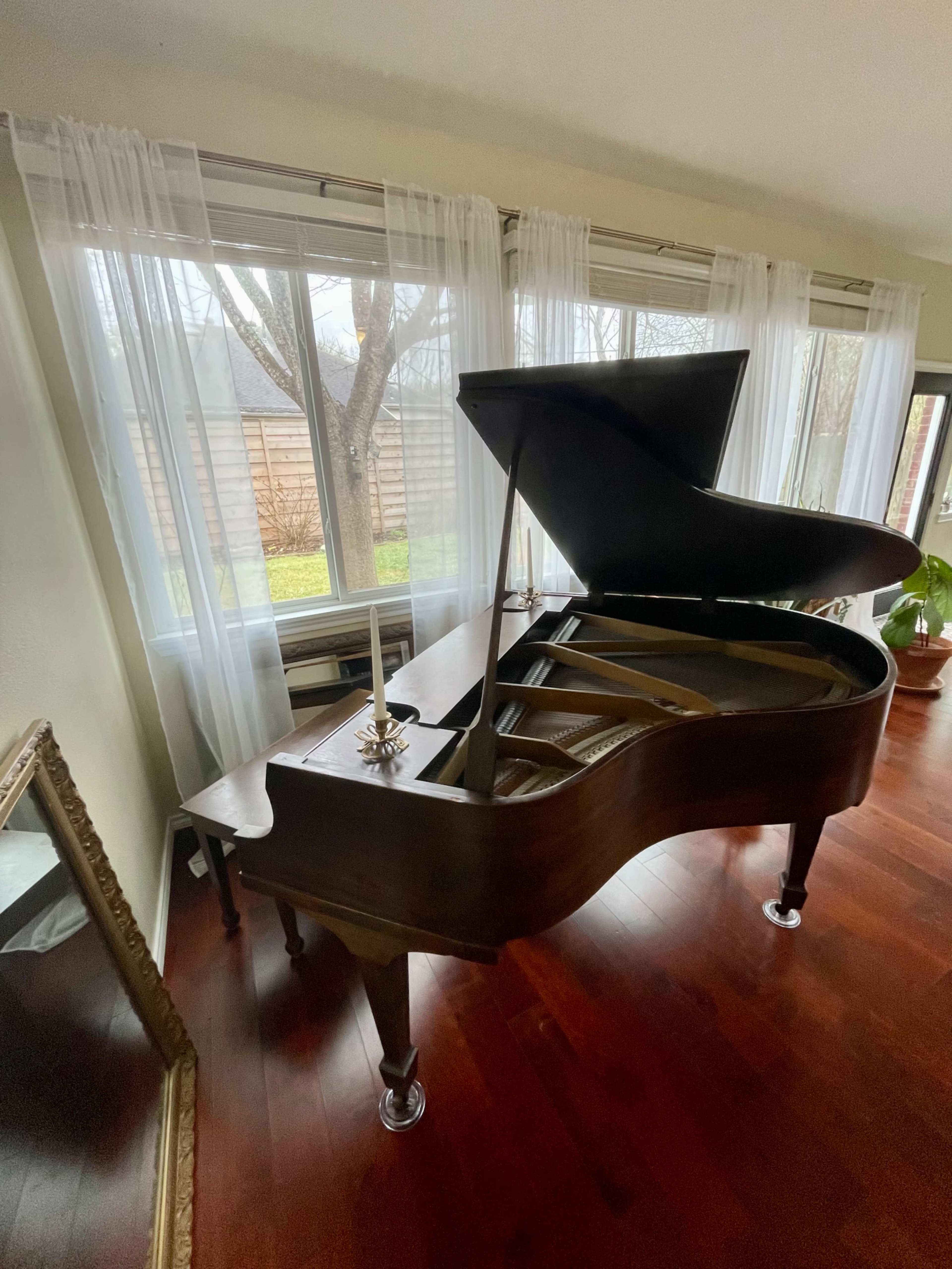 A grand piano is positioned in a sunlit room with large windows draped in sheer curtains, overlooking a garden.