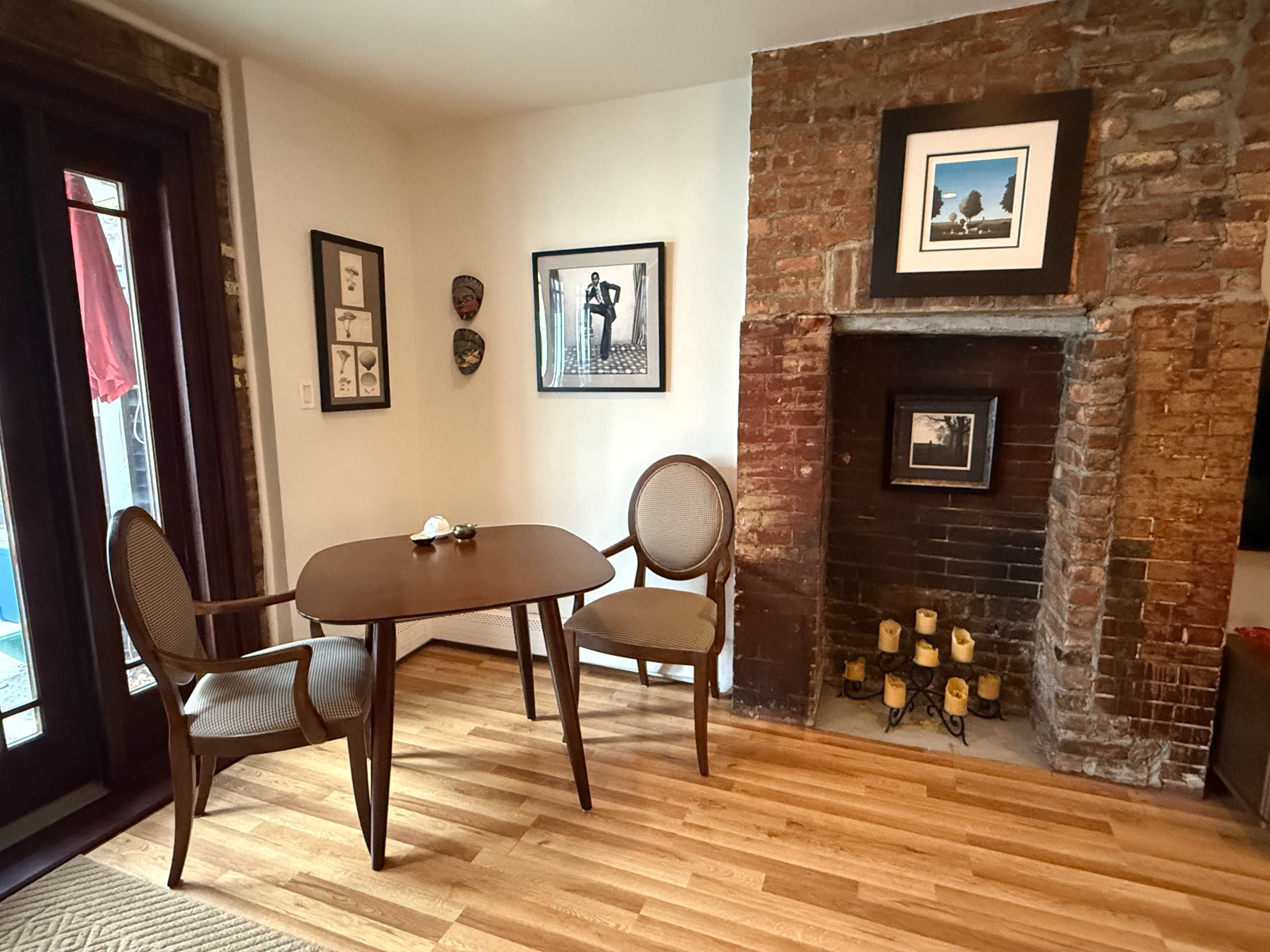 A small dining area features a round table with two chairs next to a brick fireplace, adorned with framed artwork on the walls.