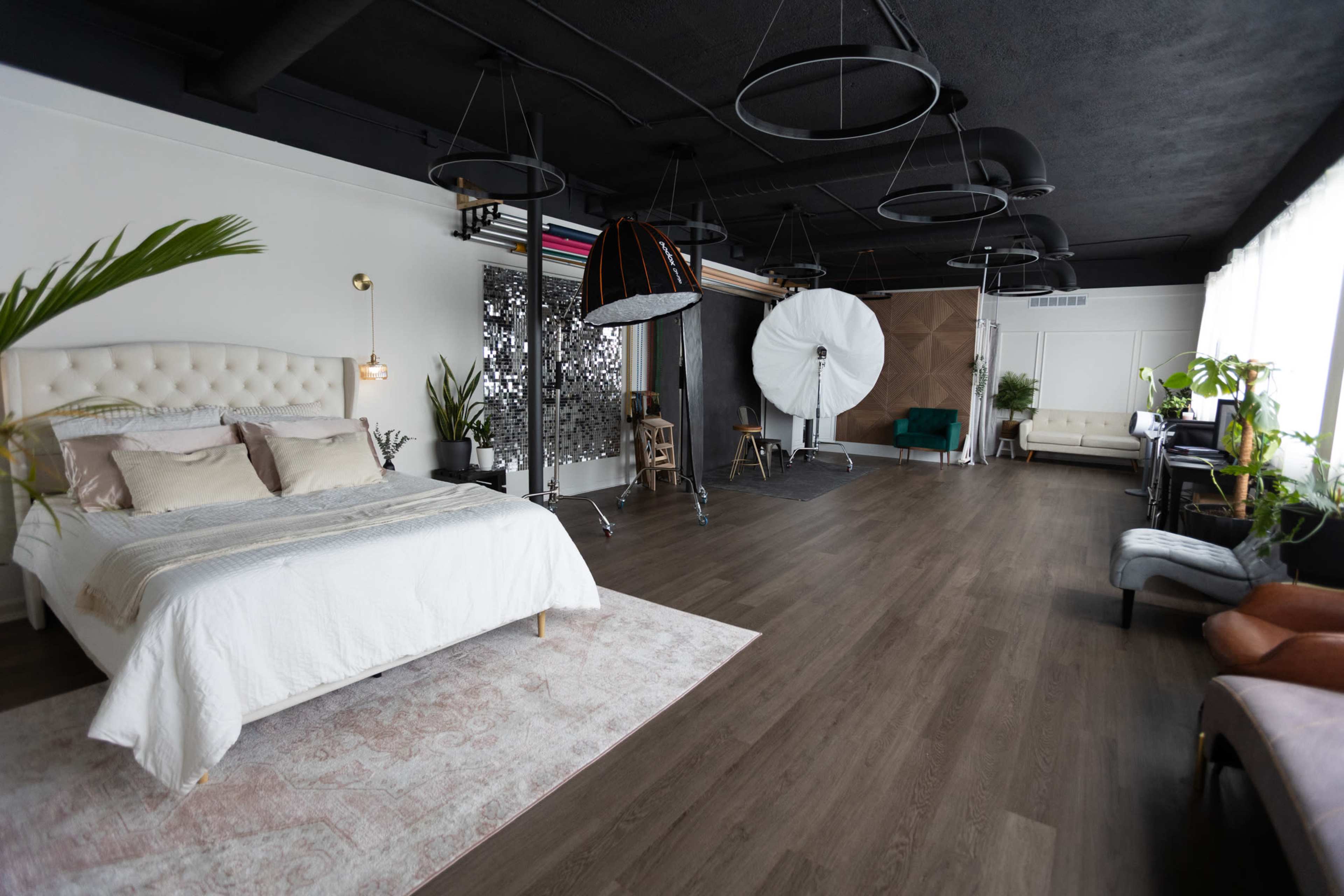 The image shows a spacious studio interior featuring a bed, various lighting equipment, and greenery, with a combination of wooden flooring and white walls.