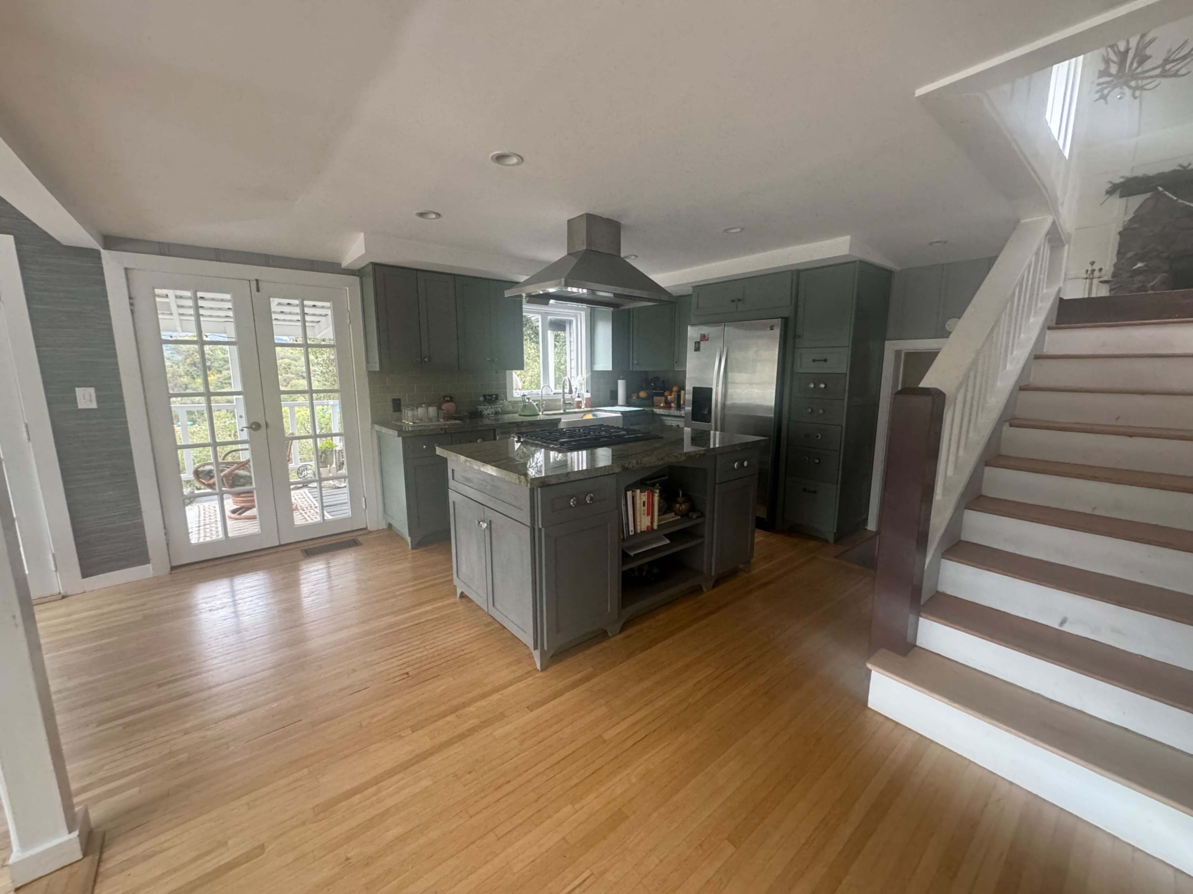 The image shows a modern kitchen with a central island, stainless steel appliances, and a staircase leading to an upper level.