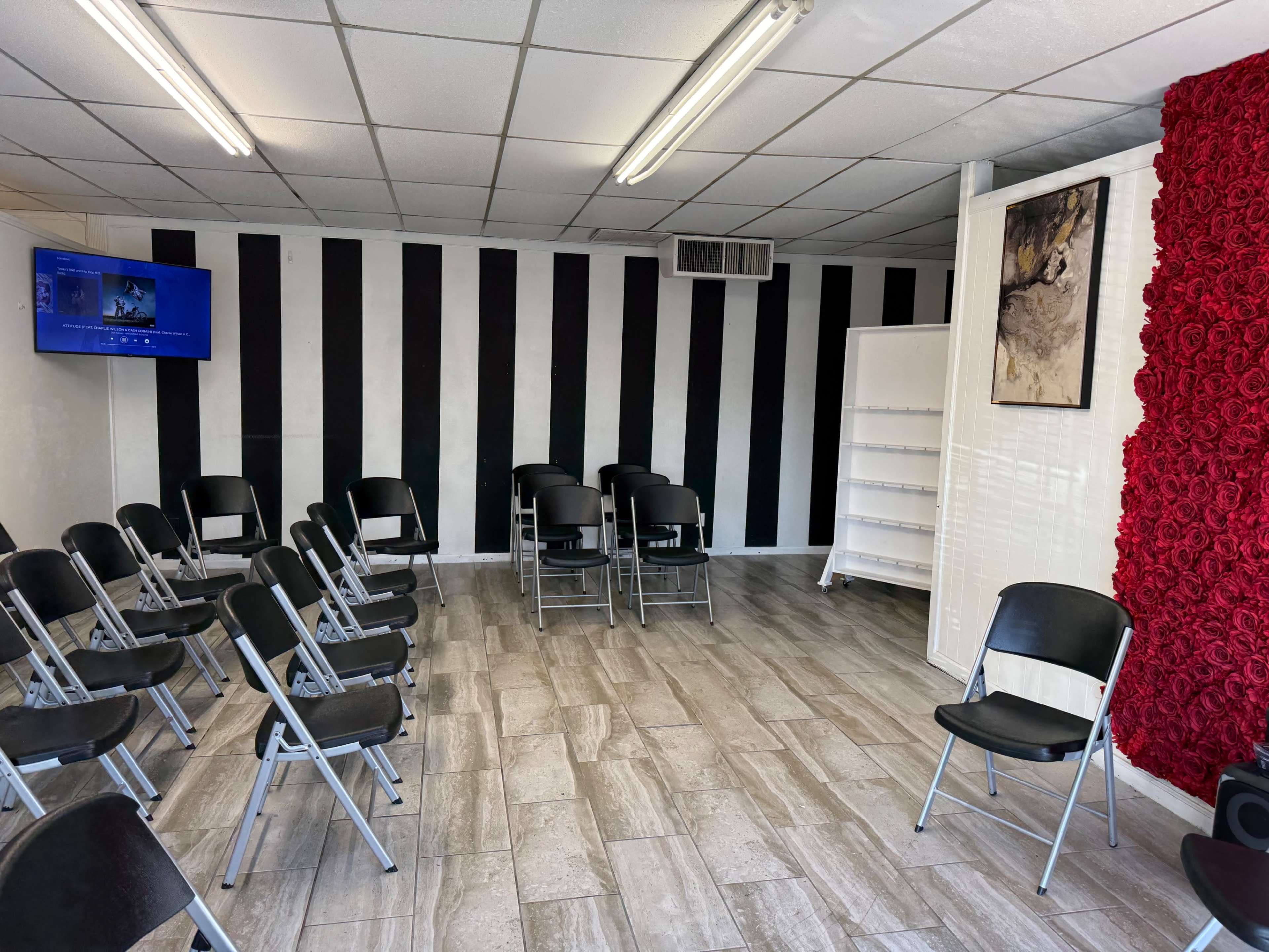 The image shows a room with black and white striped walls, empty folding chairs arranged in a semicircle, a television mounted on the wall, and a decorative wall covered in artificial red roses.