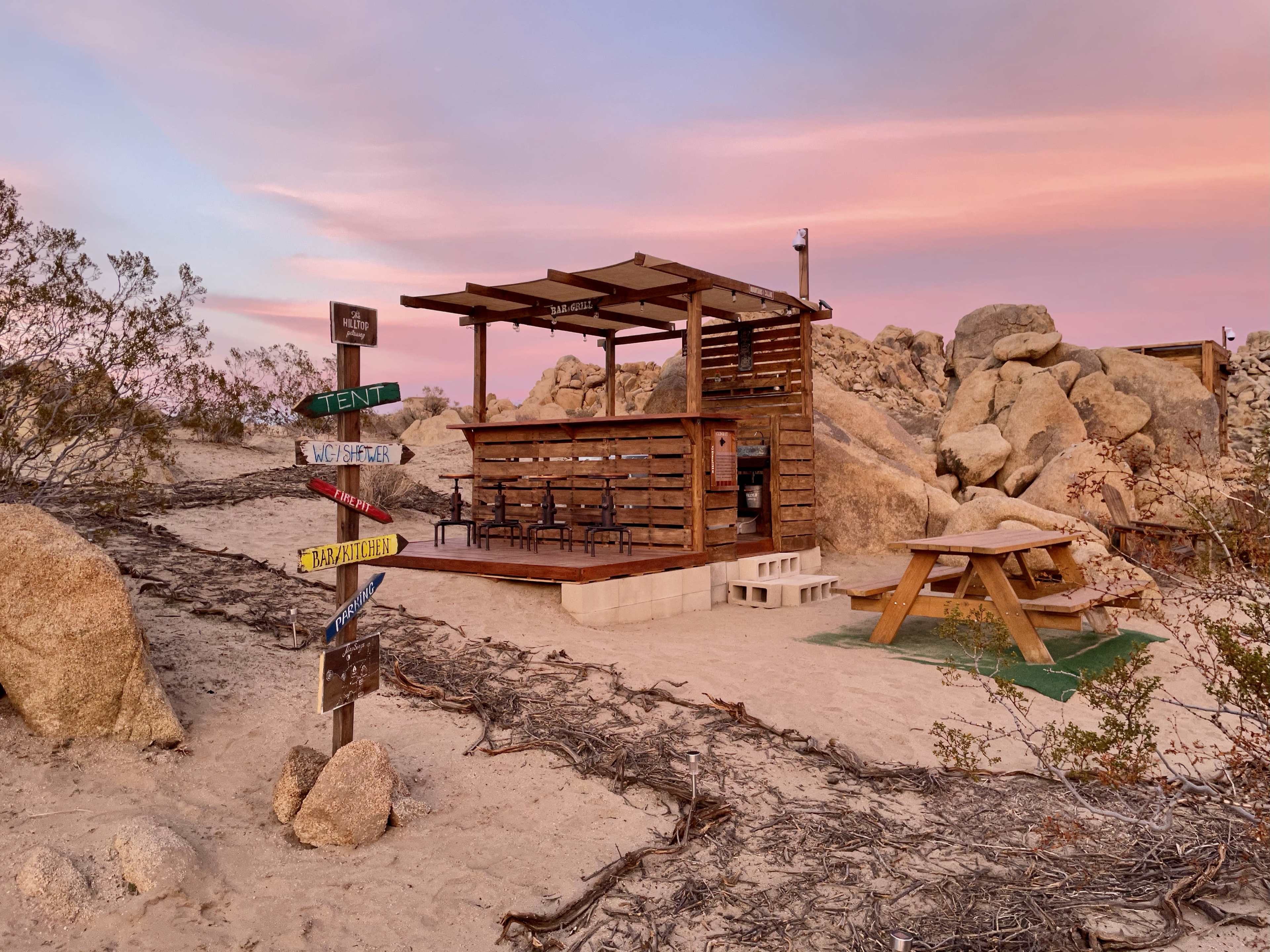 A small wooden shelter with seating options is surrounded by rocky terrain and desert vegetation under a colorful sky.