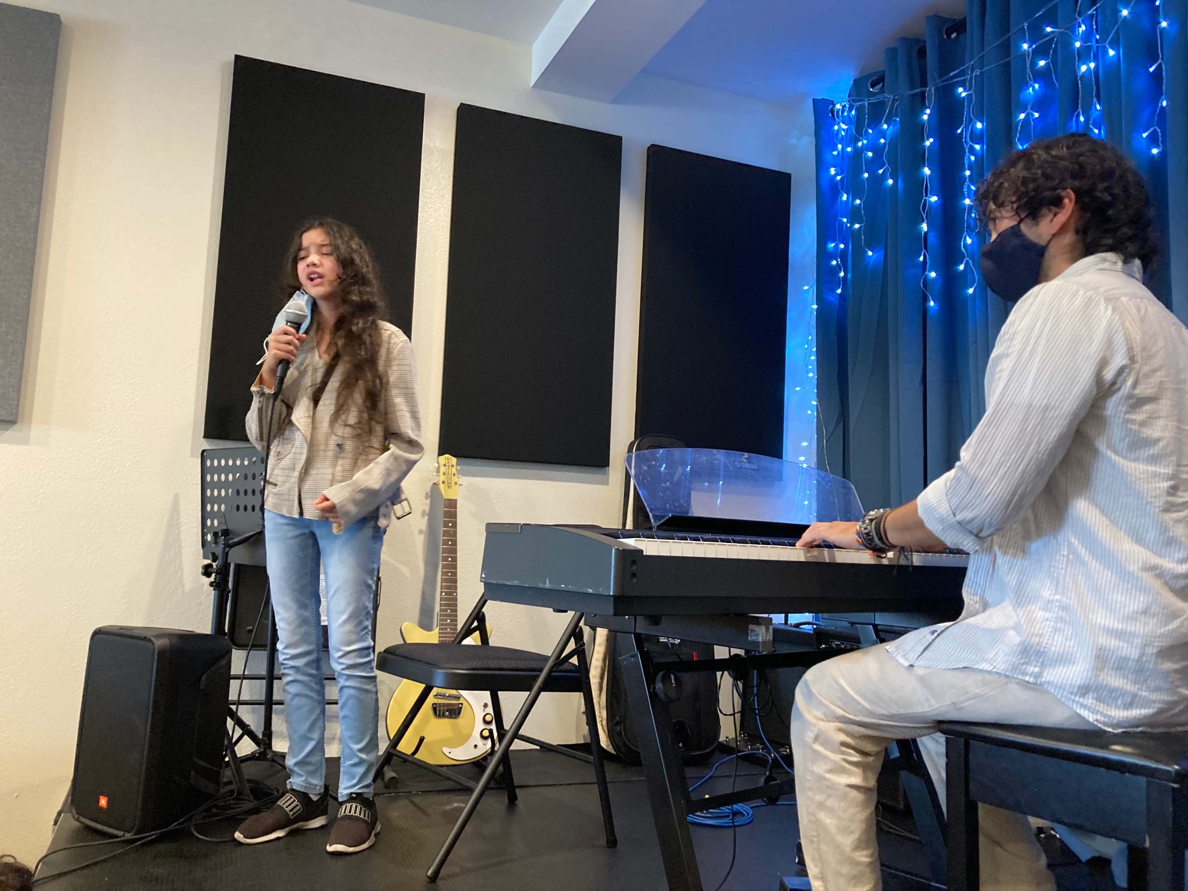 A young woman sings into a microphone while a man plays a keyboard in a room with music equipment and blue lights.