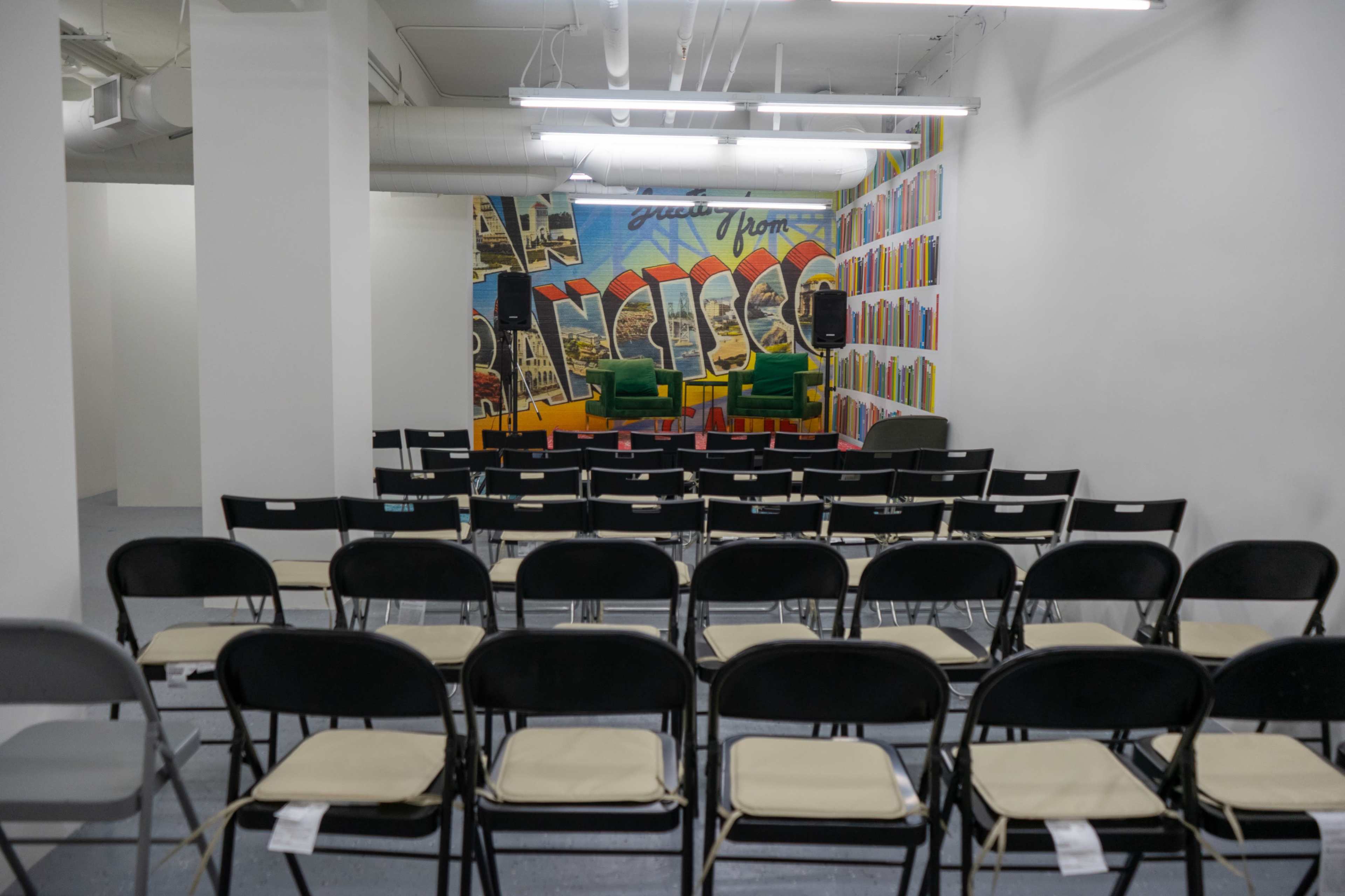 The image shows a room with rows of black folding chairs facing a mural featuring colorful book spines and the phrase "Greetings from San Francisco."