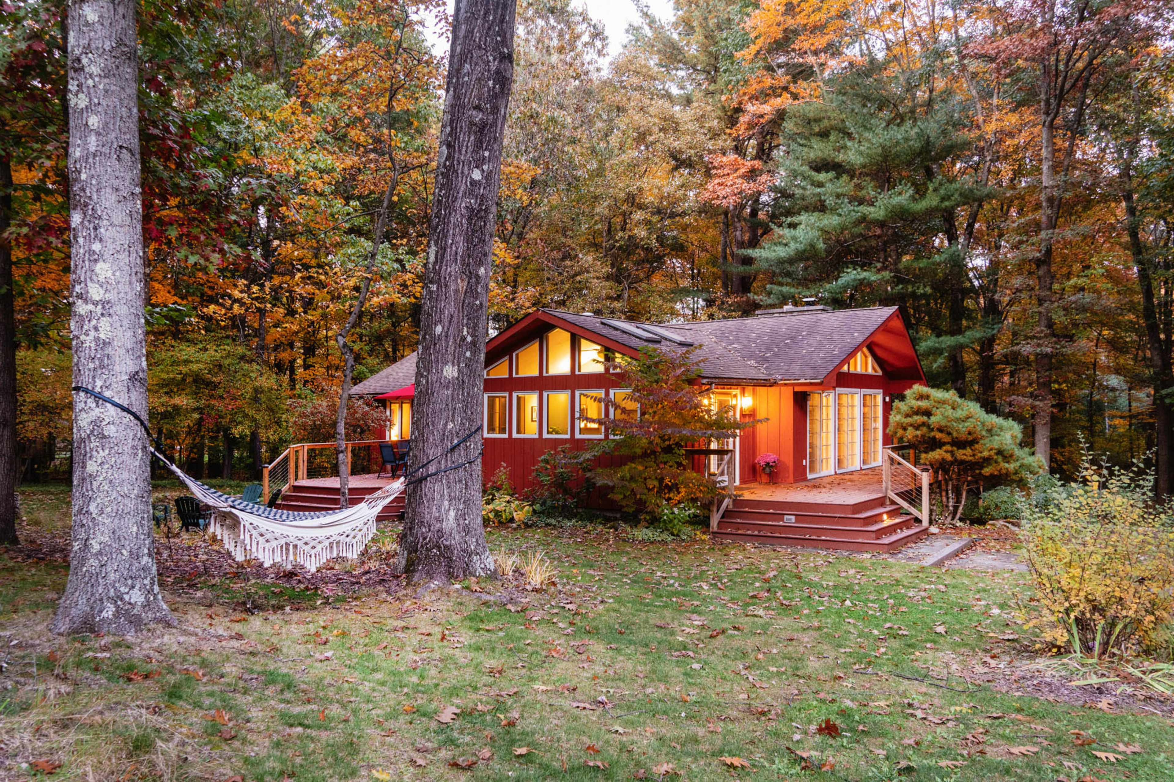A red cabin with large windows is surrounded by trees and autumn foliage, featuring a hammock tied between two trees in the foreground.