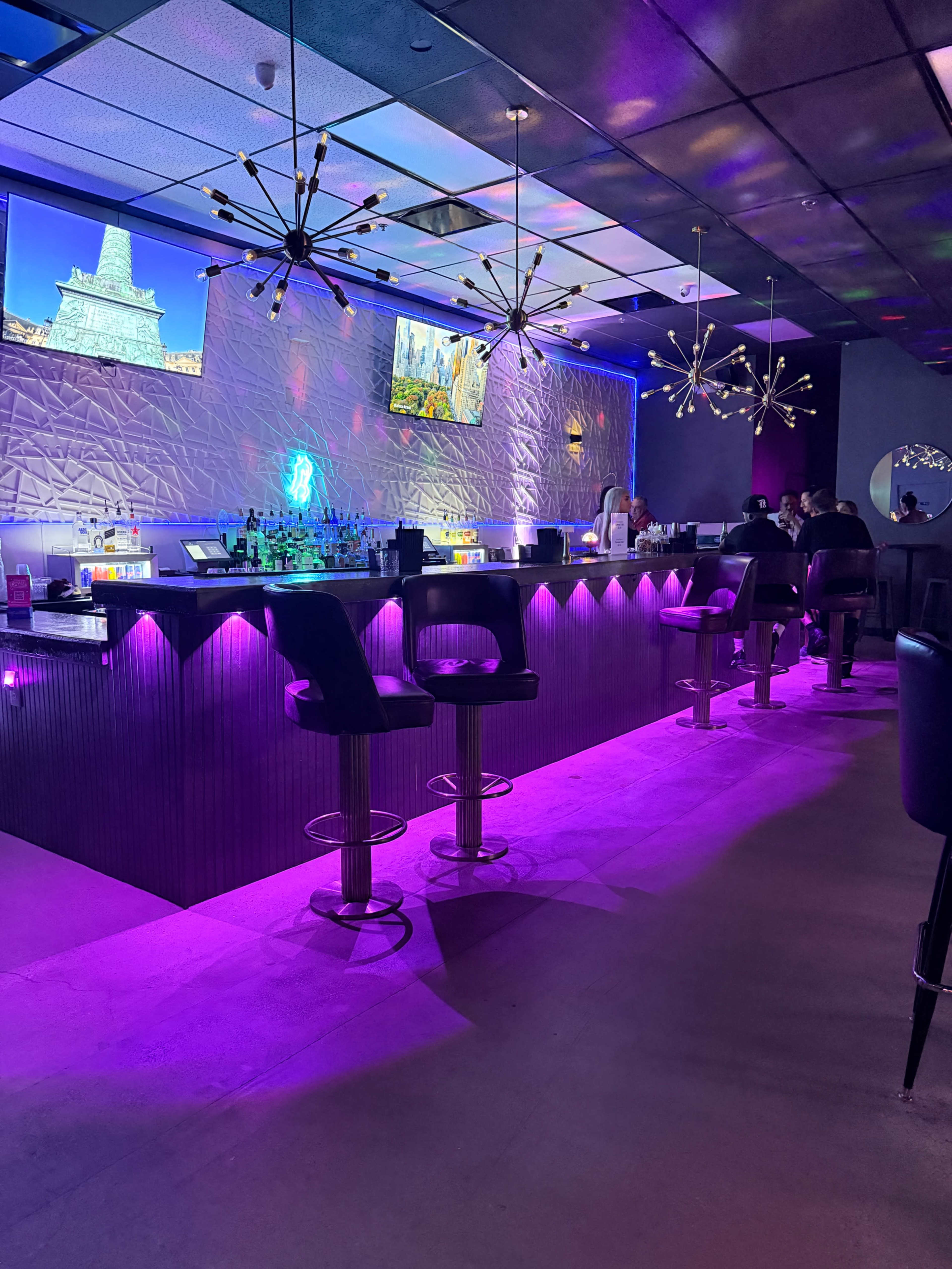 A modern bar with a glowing purple backlit counter, two televisions displaying images, and patrons seated at high stools.