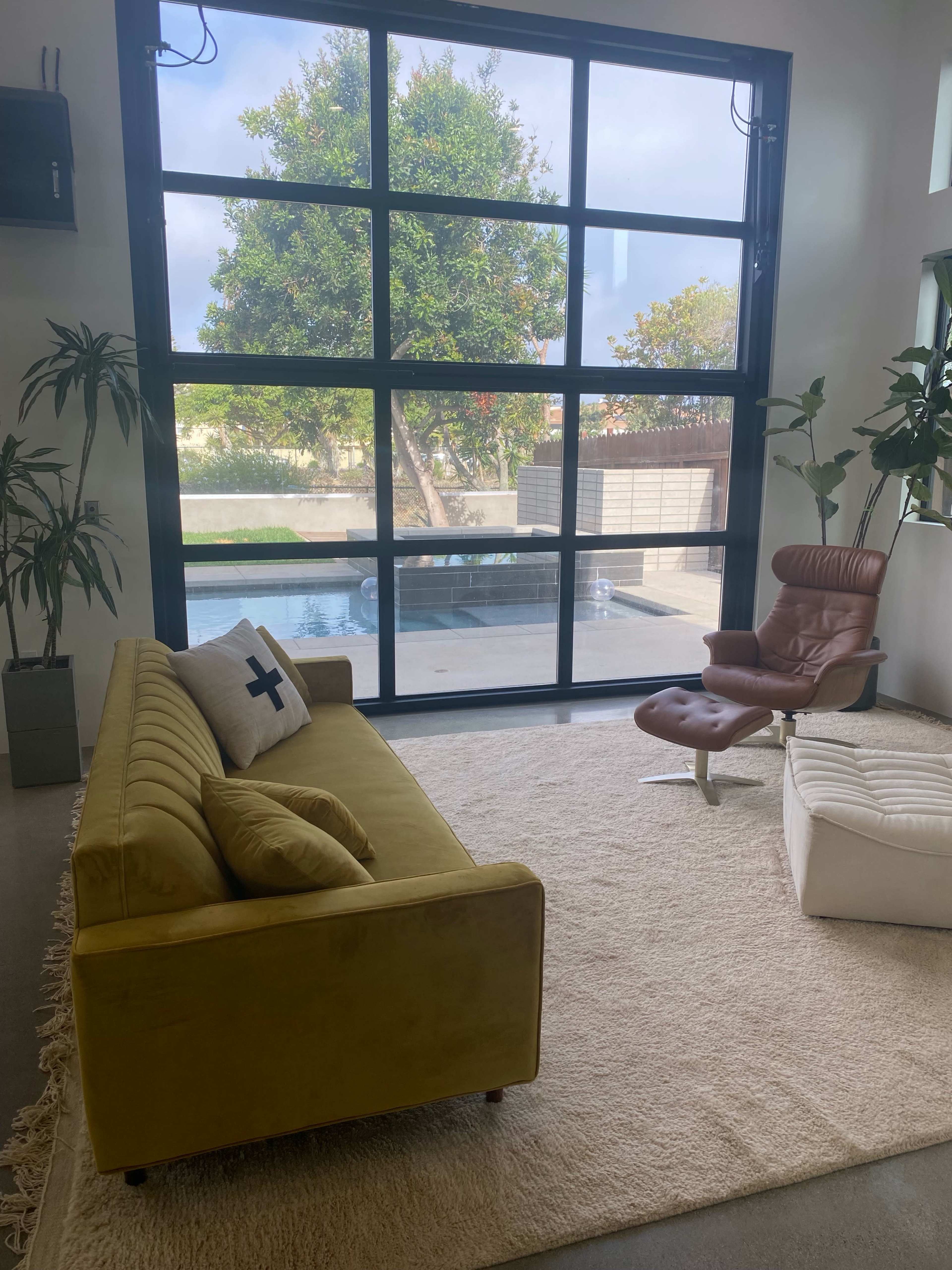 A modern living room features a yellow sofa, a brown armchair, and large windows overlooking a pool and garden.