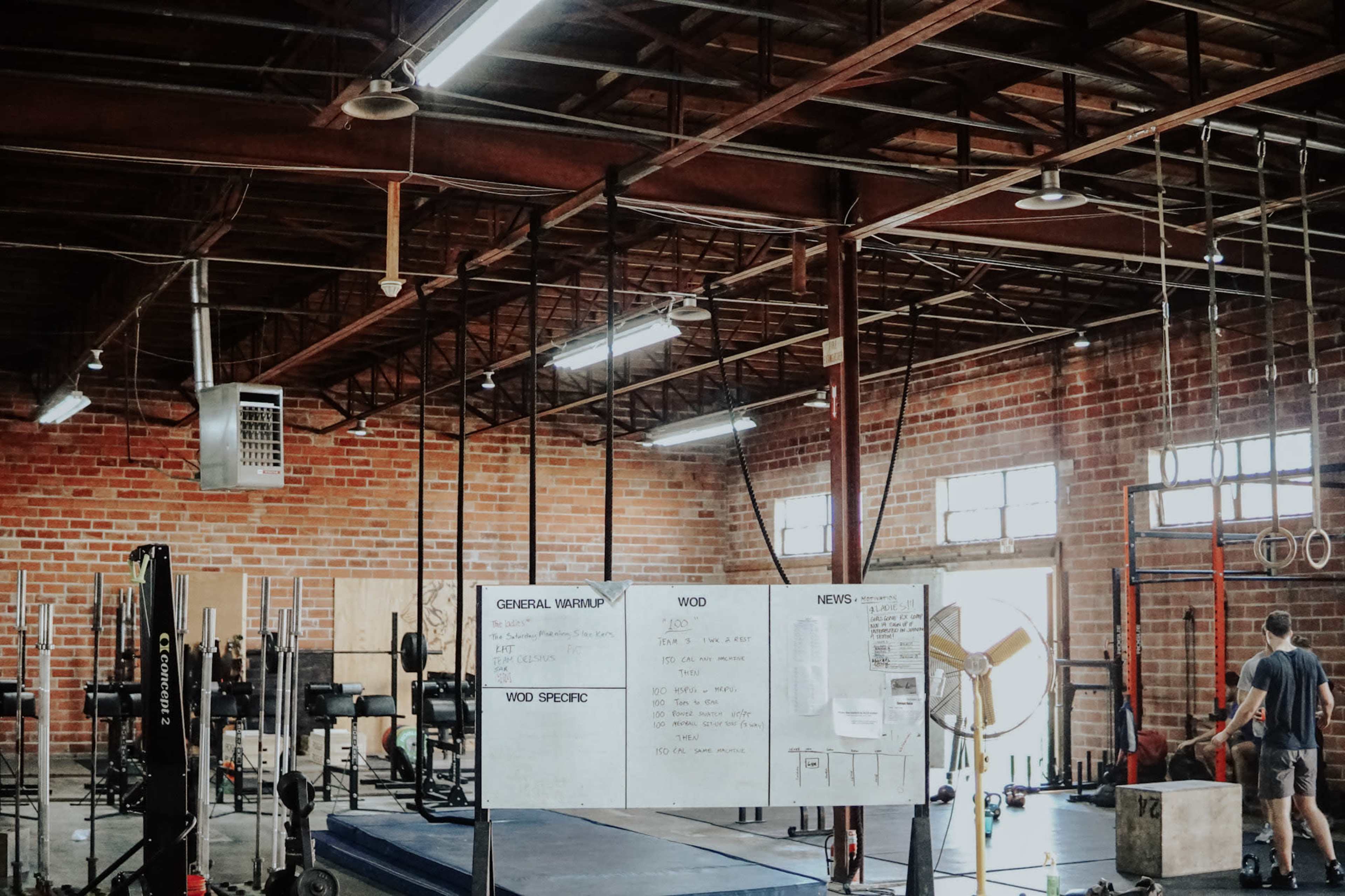 A spacious gym with brick walls features various weightlifting equipment, a large whiteboard for workout instructions, and several participants engaged in exercises.