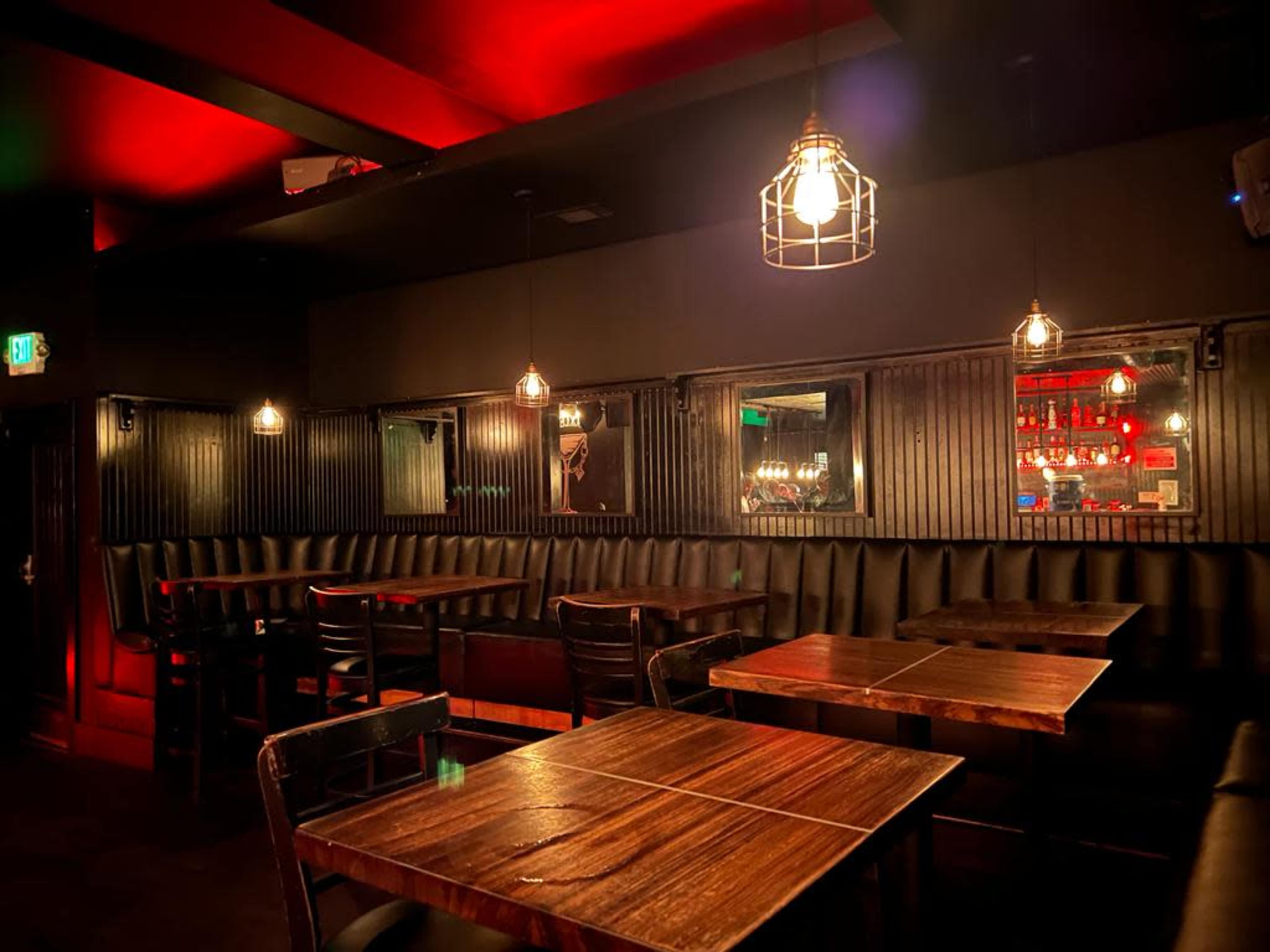 A dimly lit bar interior with wooden tables, black leather seating, and hanging pendant lights.